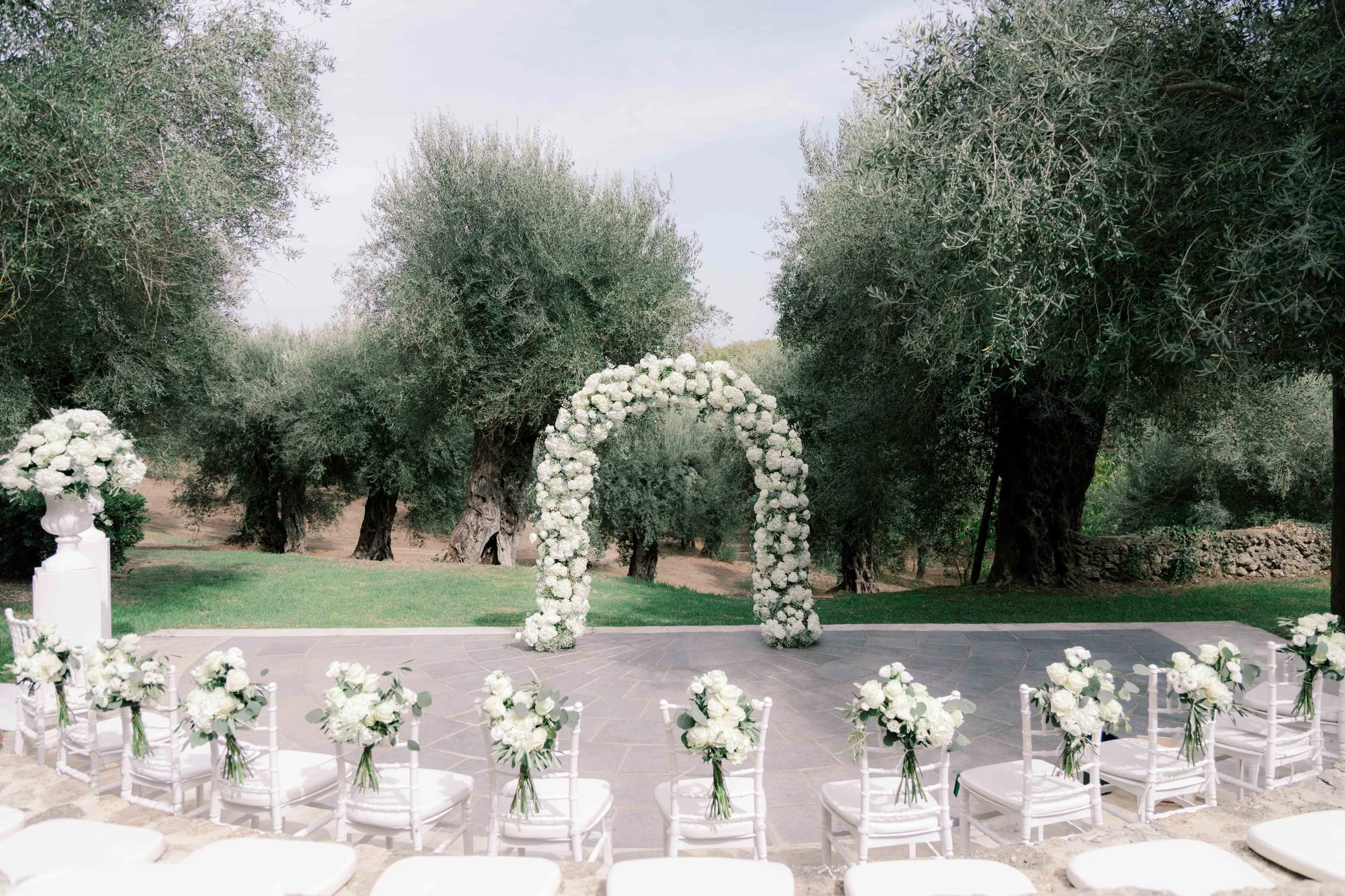 Empty outdoor wedding ceremony setup with white chairs decorated with white flowers, a floral arch, and trees in the background.