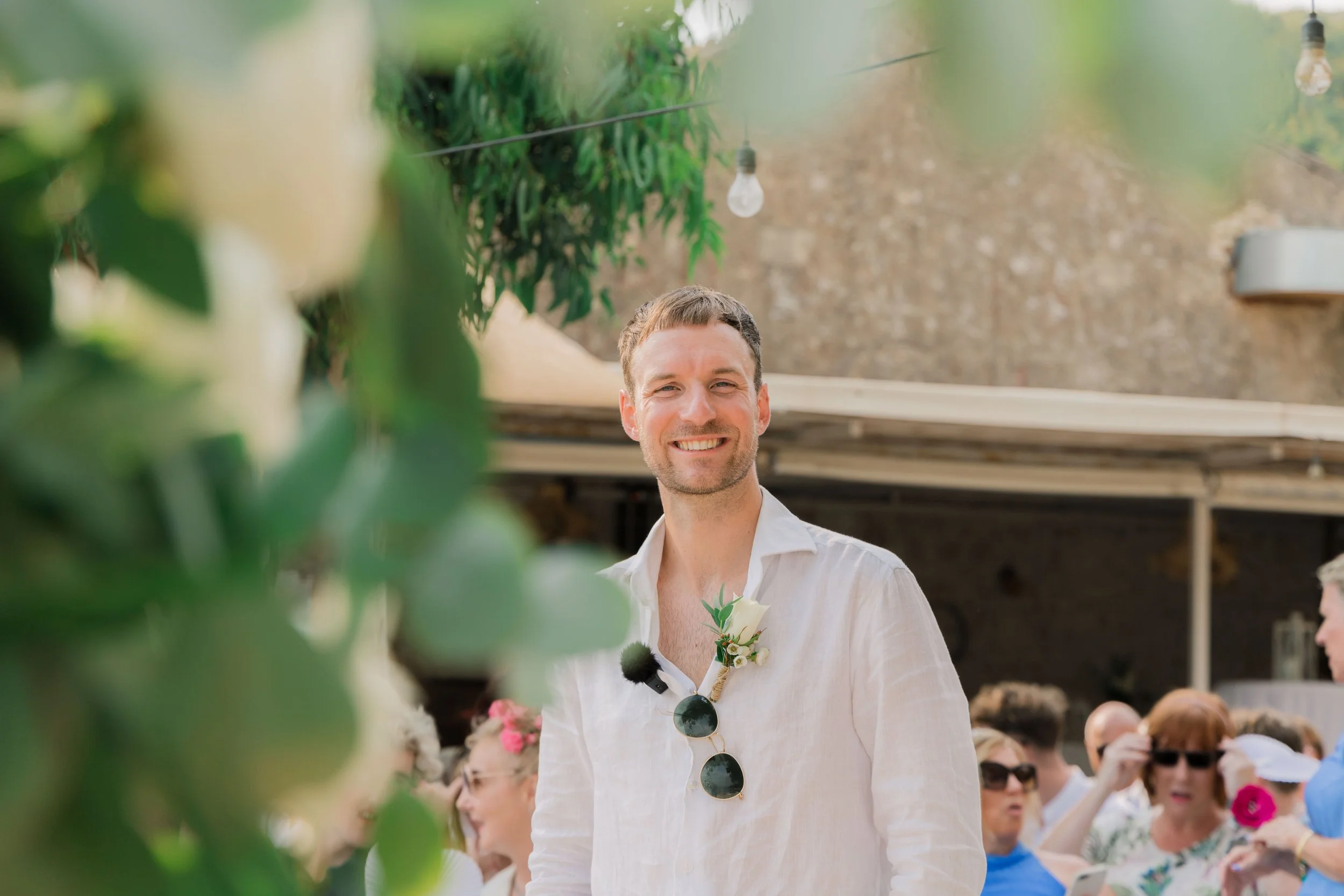 A smiling man with short brown hair and beard, wearing a white linen shirt and sunglasses hanging on the collar, standing outdoors at a social gathering during daytime, with people and green foliage in the background.