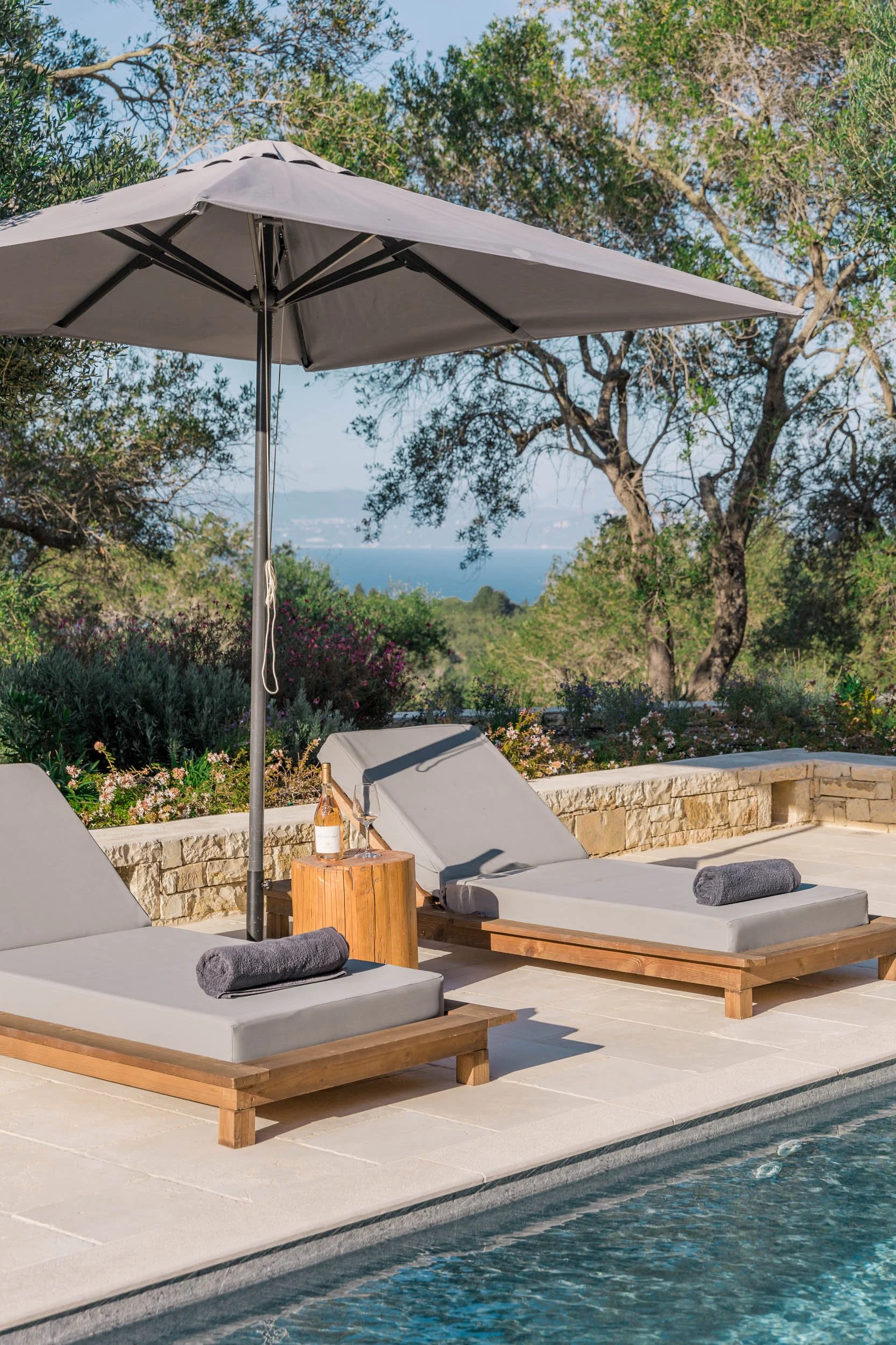 Poolside scene with two lounge chairs, gray cushions, and rolled towels, under a large grey umbrella, with a stone wall, trees, and a view of water in the background.