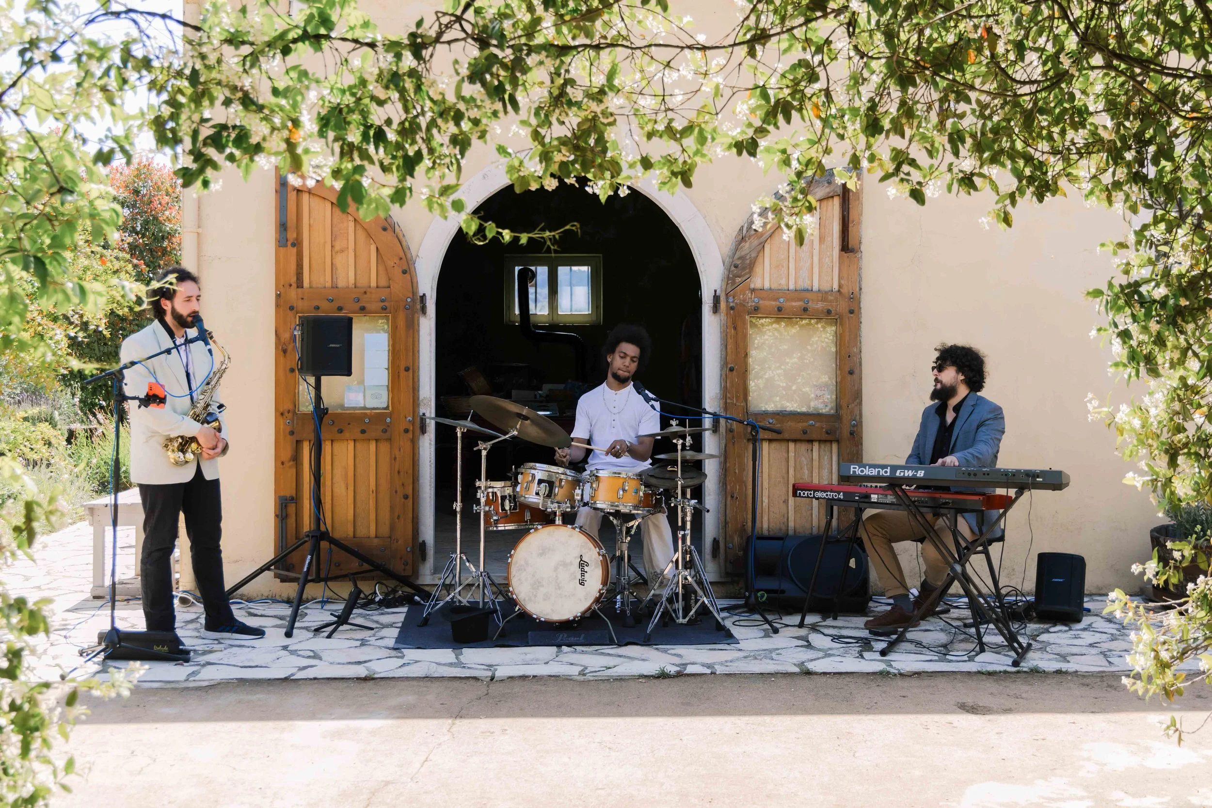 Three musicians performing outside a building with wooden doors, a saxophonist on the left, a drummer in the center, and a keyboard player on the right, under a tree with green foliage.