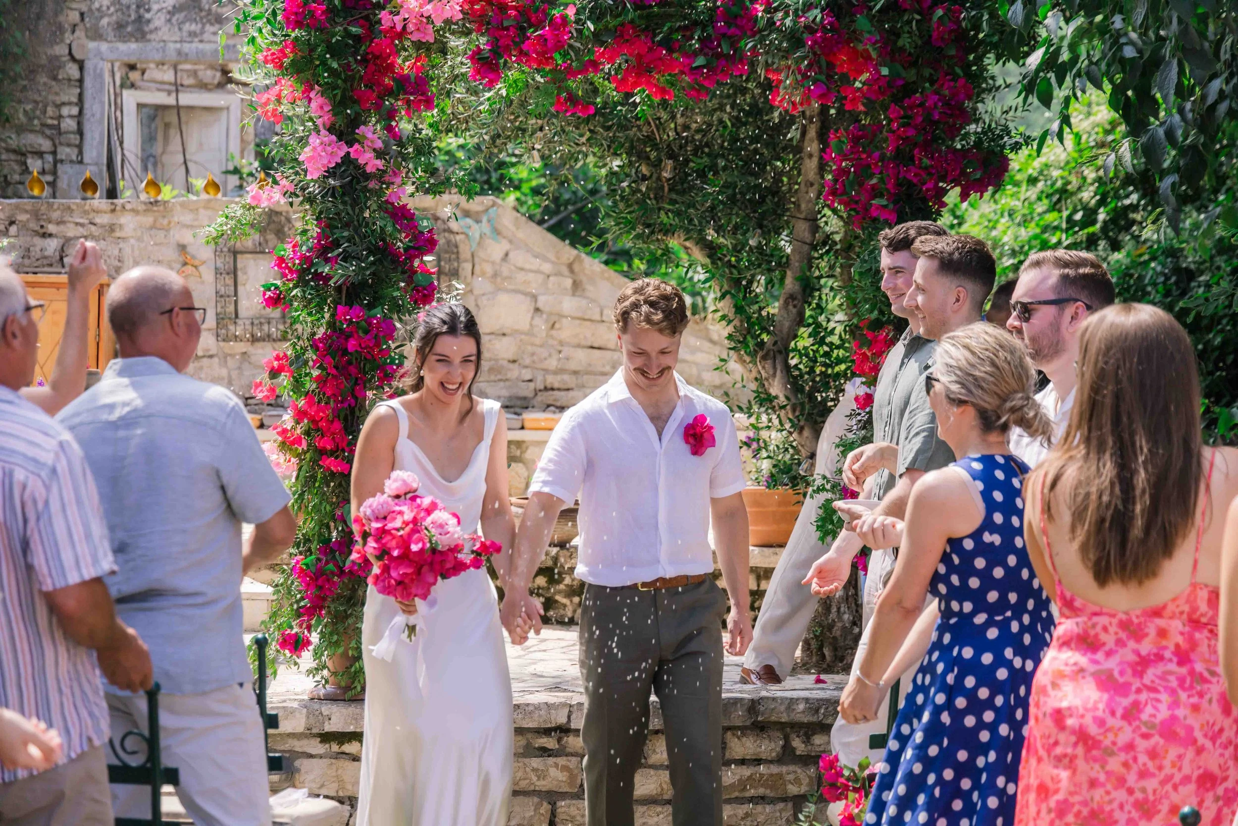 A wedding celebration outdoors with a bride and groom holding hands under a floral arch, surrounded by family and friends, with bright pink and purple flowers and green foliage in the background.