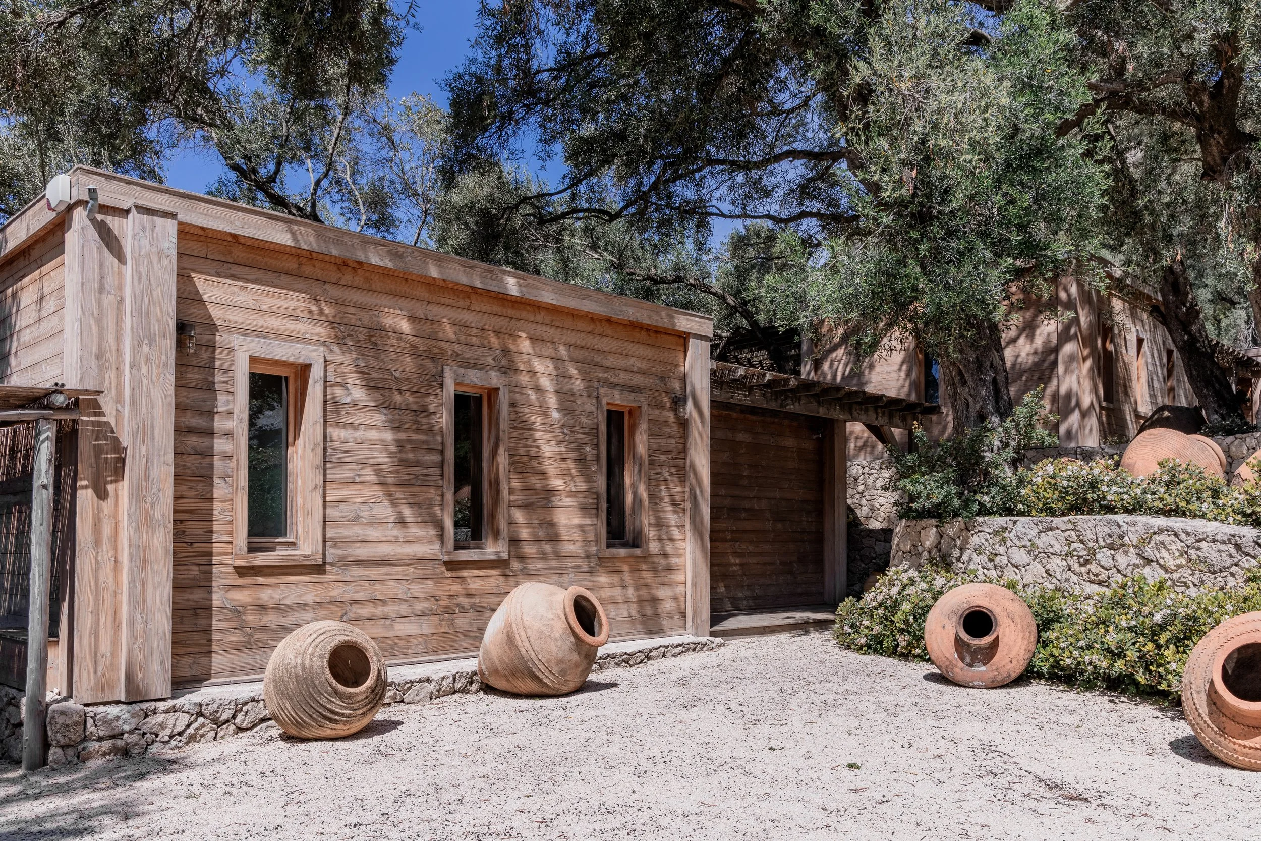 A rustic wooden building with three tall, narrow windows, surrounded by greenery and large clay pots on a gravel surface, under a clear blue sky.
