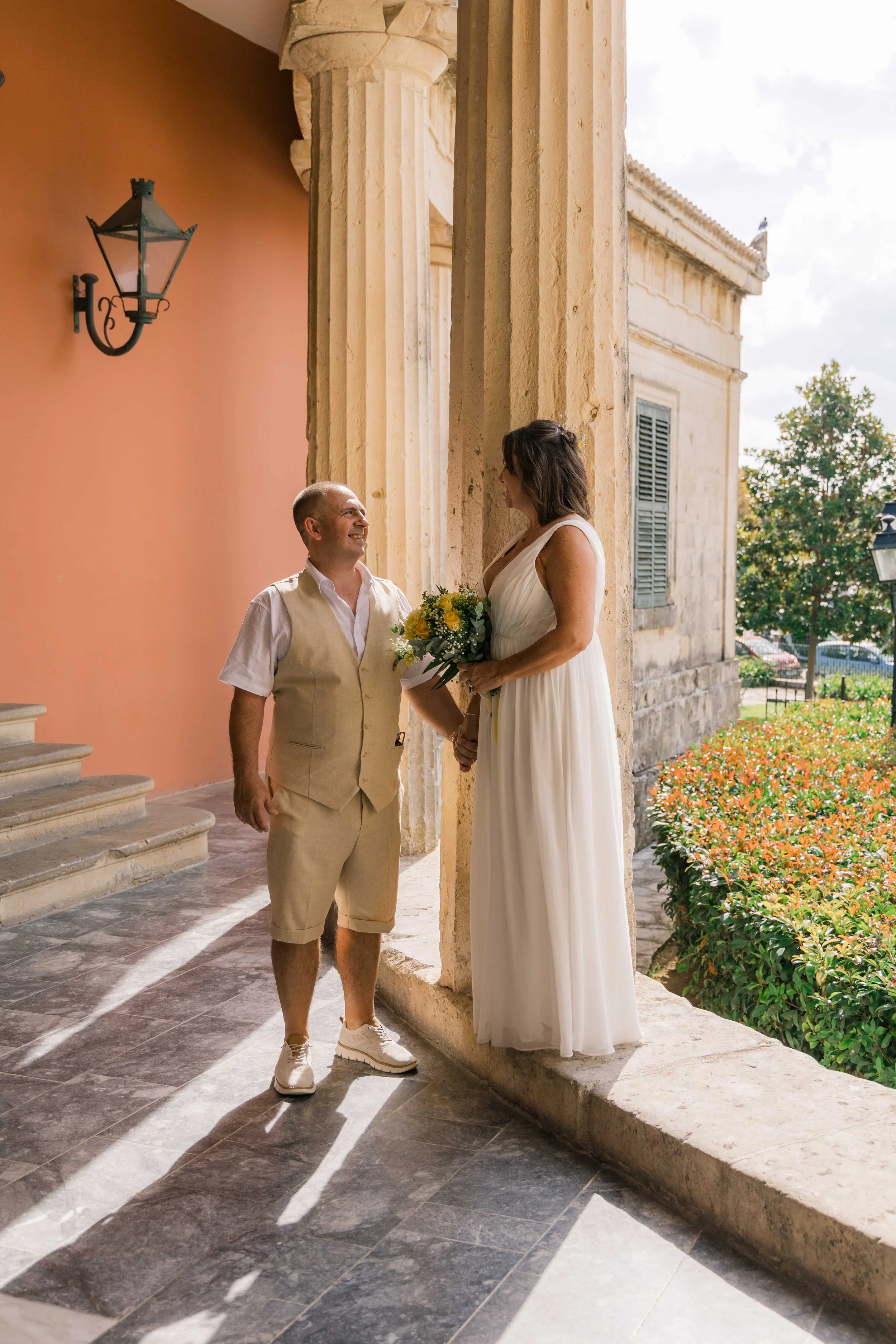 A man and woman are standing outside a historic building having a conversation. The woman is holding a bouquet of yellow flowers and wearing a white dress, while the man is dressed in a light beige vest and shorts. Sunlight casts long shadows on the 