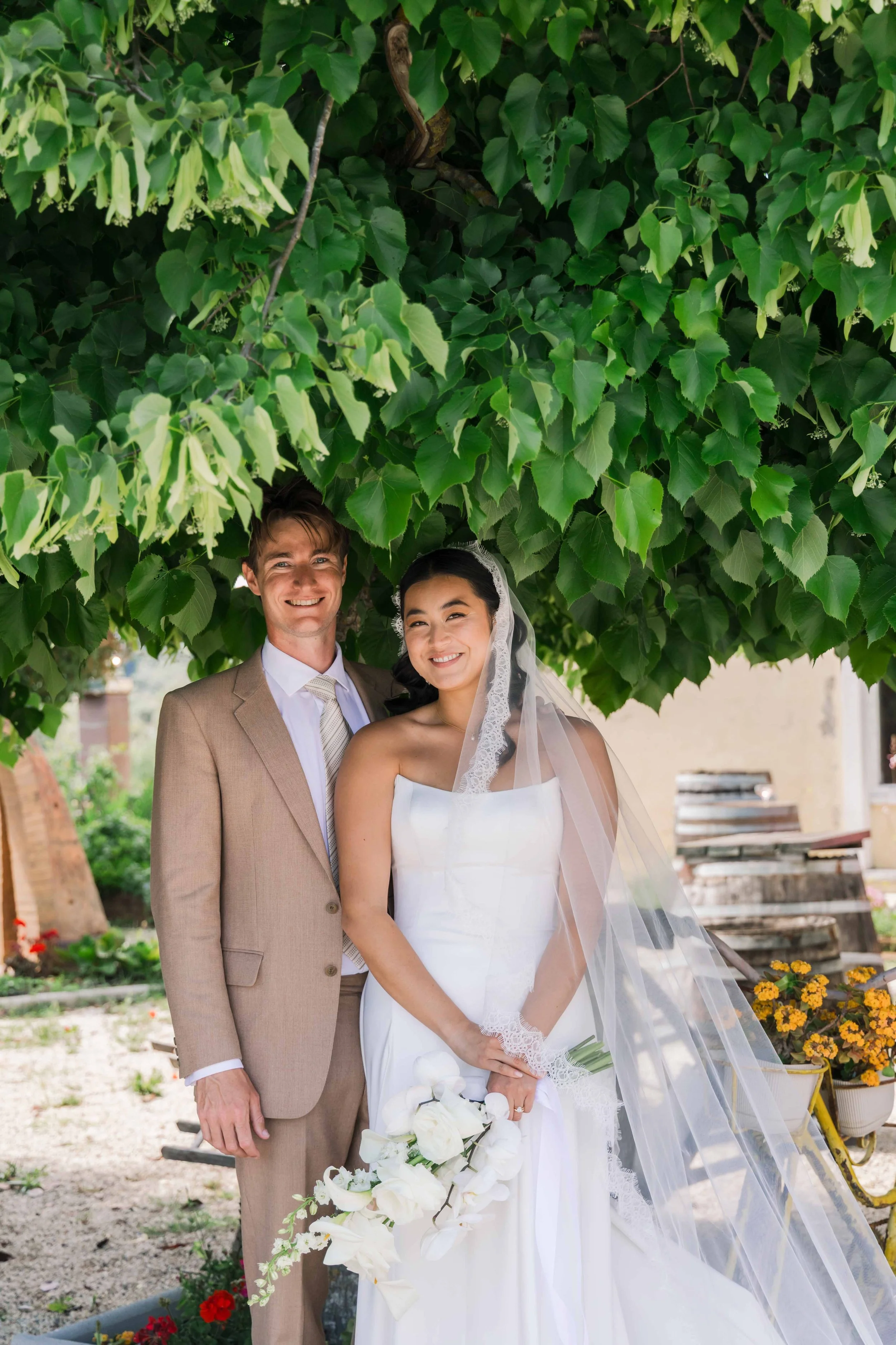 A smiling bride and groom standing outdoors beneath a lush green tree, with the bride holding a white flower bouquet.