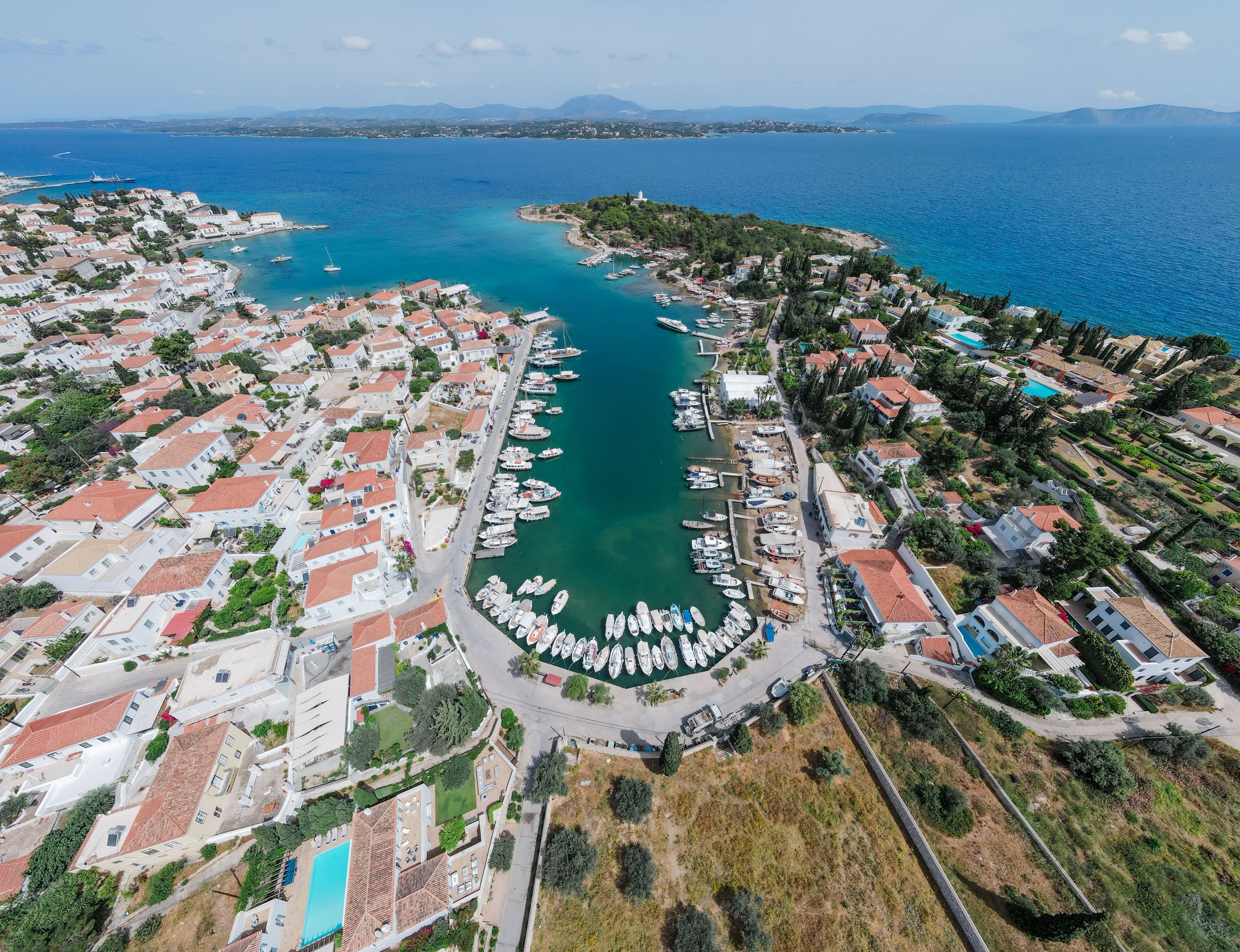Aerial view of a seaside town with a marina filled with boats, red-roofed houses, and surrounding green hills and ocean.
