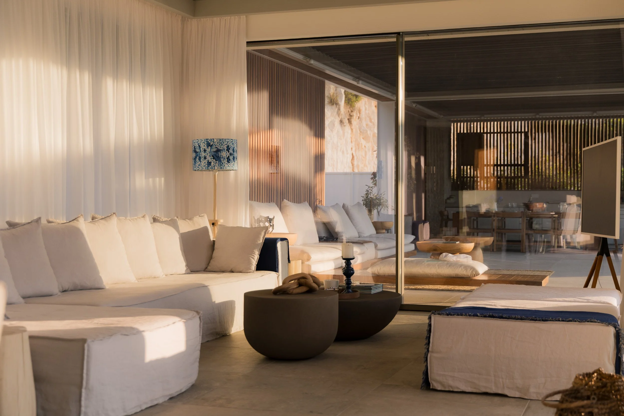 Modern living room with white sofa, beige cushions, black and beige coffee tables, standing lamp with blue patterned shade, and large glass sliding doors revealing an outdoor patio with dining area.