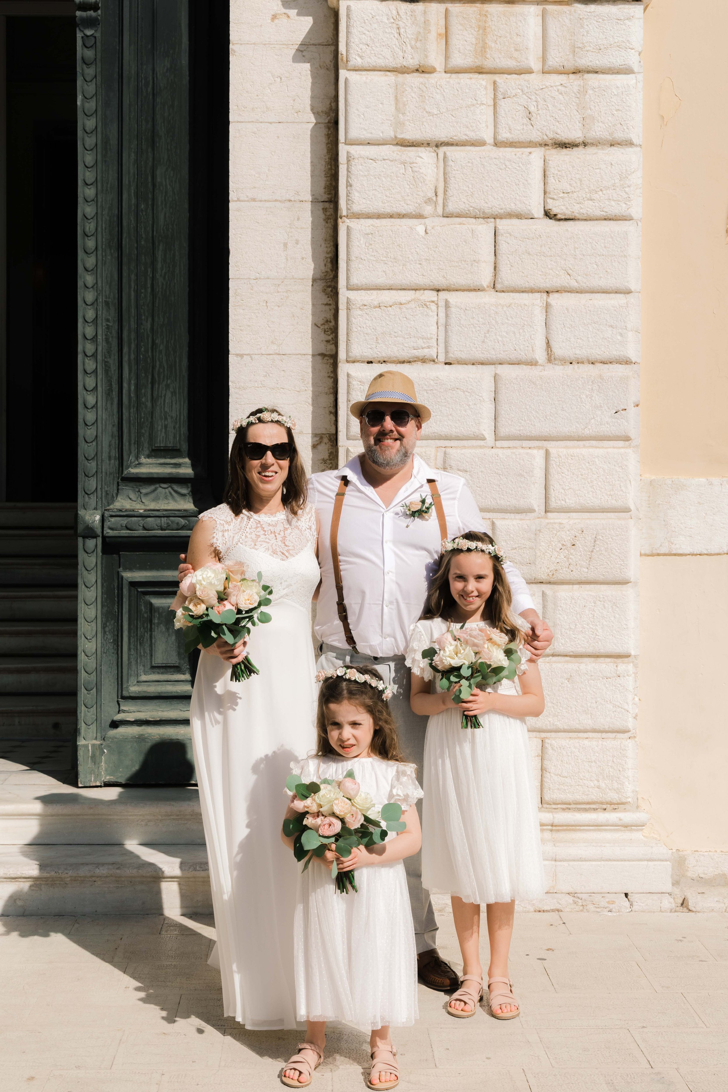 A family of five at a wedding, standing outdoors near a brick building and a large green door. The bride and two young girls are in white dresses holding bouquets of flowers, and the groom is in a white shirt with suspenders and a hat.