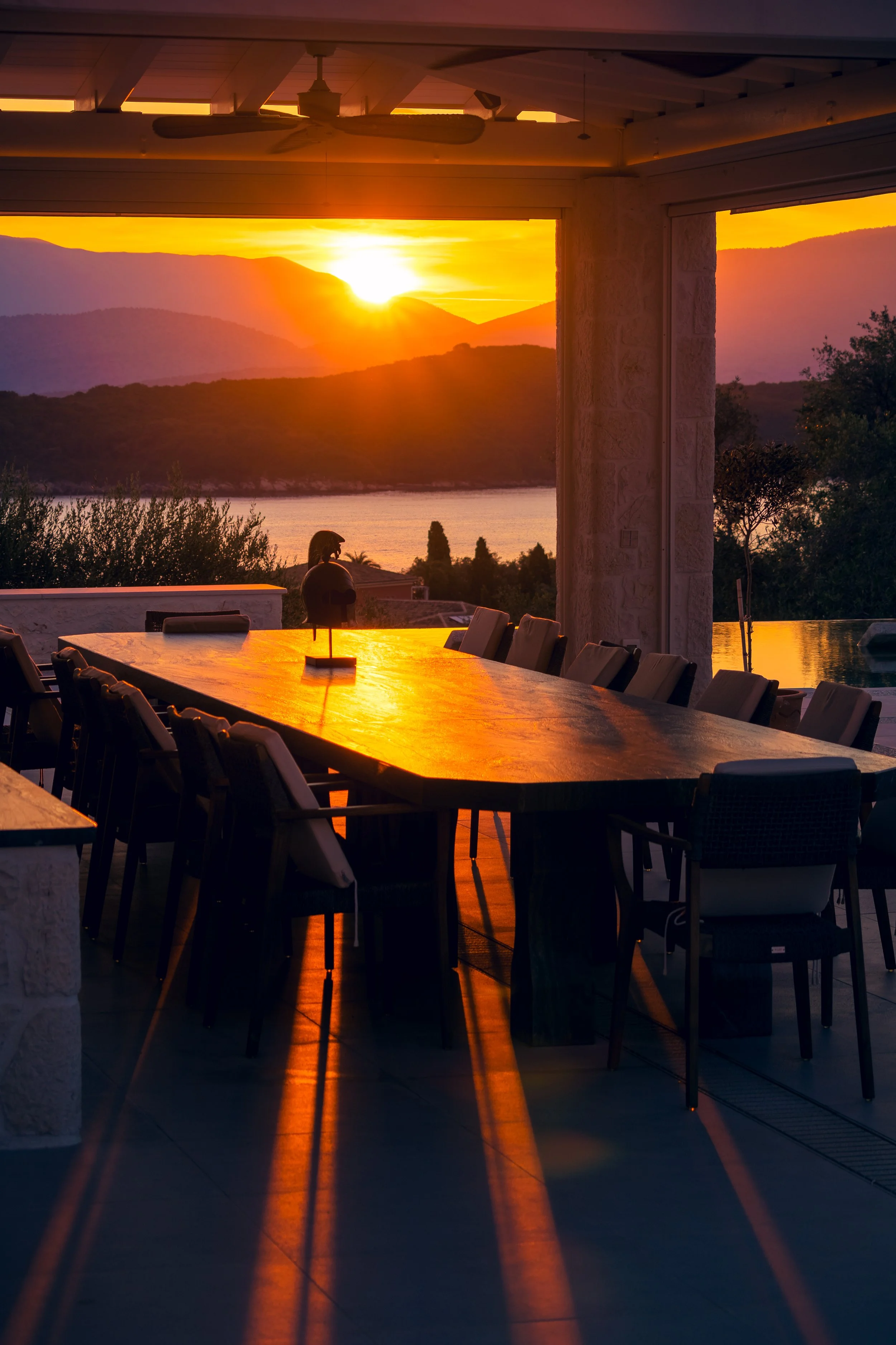 Indoor dining area with a large wooden table and chairs, overlooking a scenic sunset view over mountains and water outside, with the sun casting long shadows.