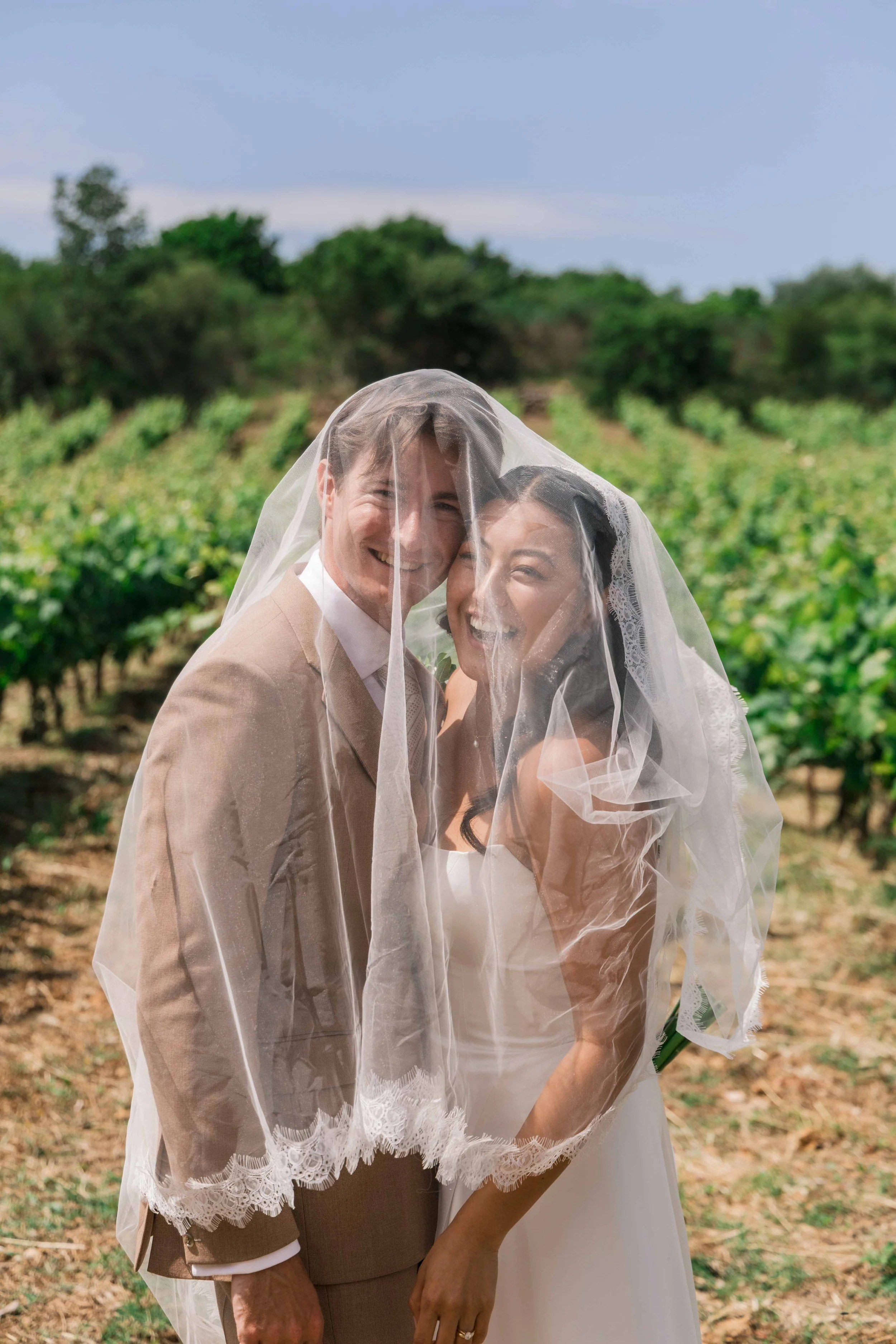 A newlywed couple, a man in a beige suit and a woman in a white wedding dress, sharing a joyful moment under a veil in a vineyard with lush green rows of grapevines in the background.