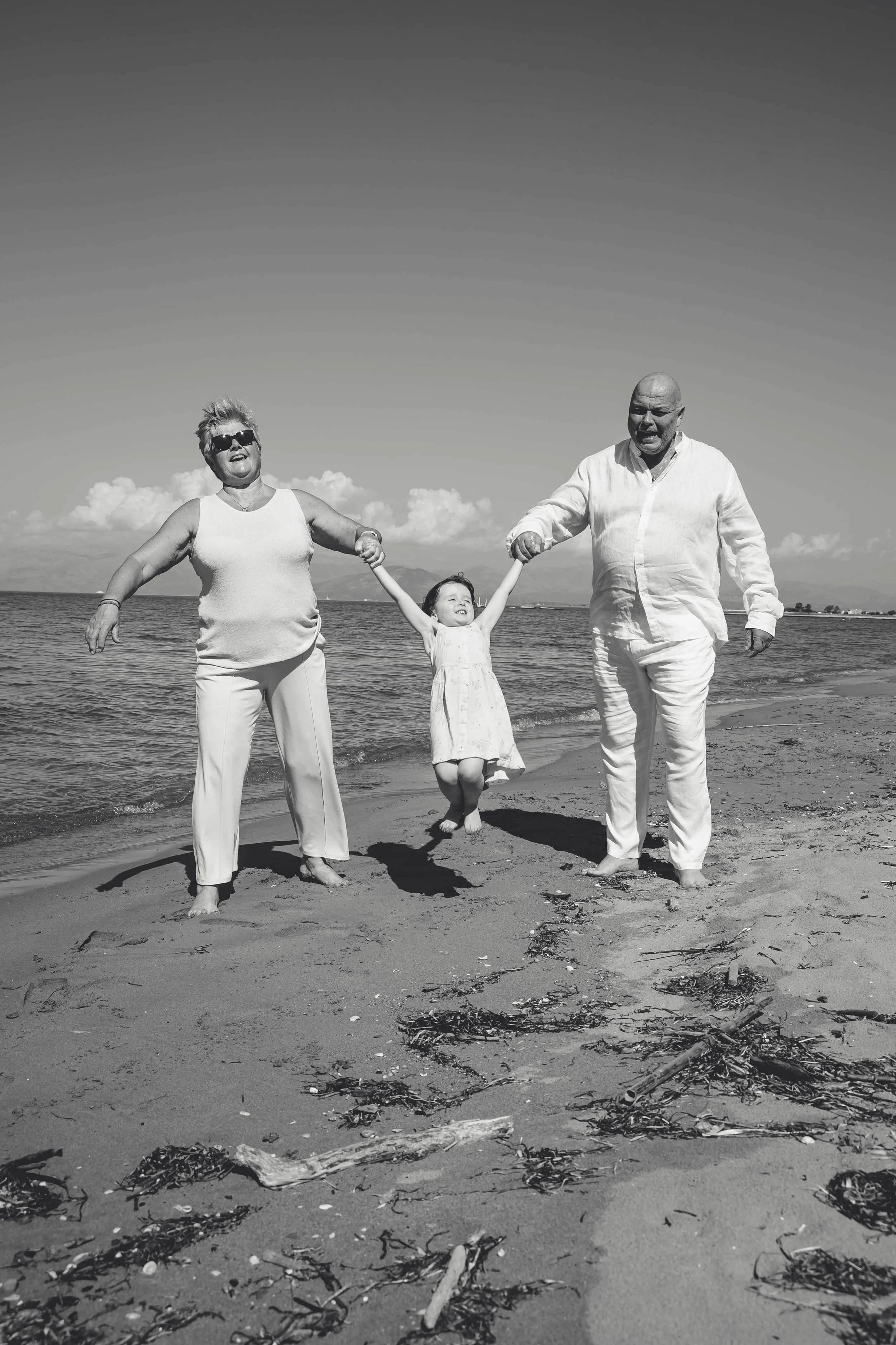 Three people holding hands and jumping on the beach, with water and clouds in the background.