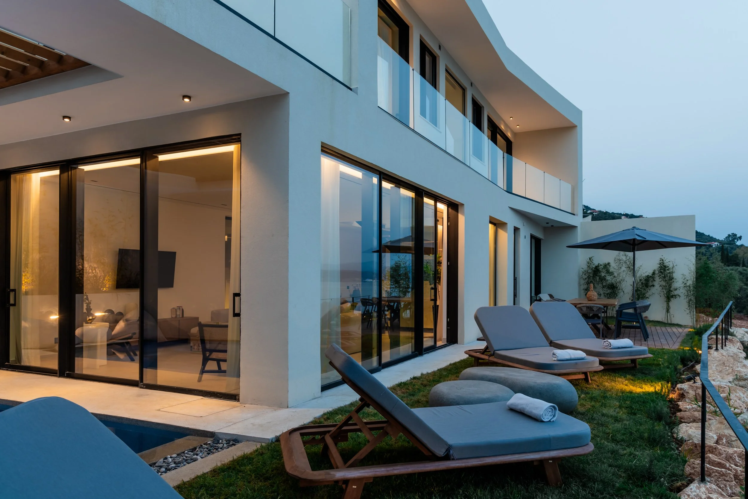 Modern white multi-story house with large glass sliding doors leading to a backyard patio with lounge chairs, a table with an umbrella, and landscaped greenery during evening.