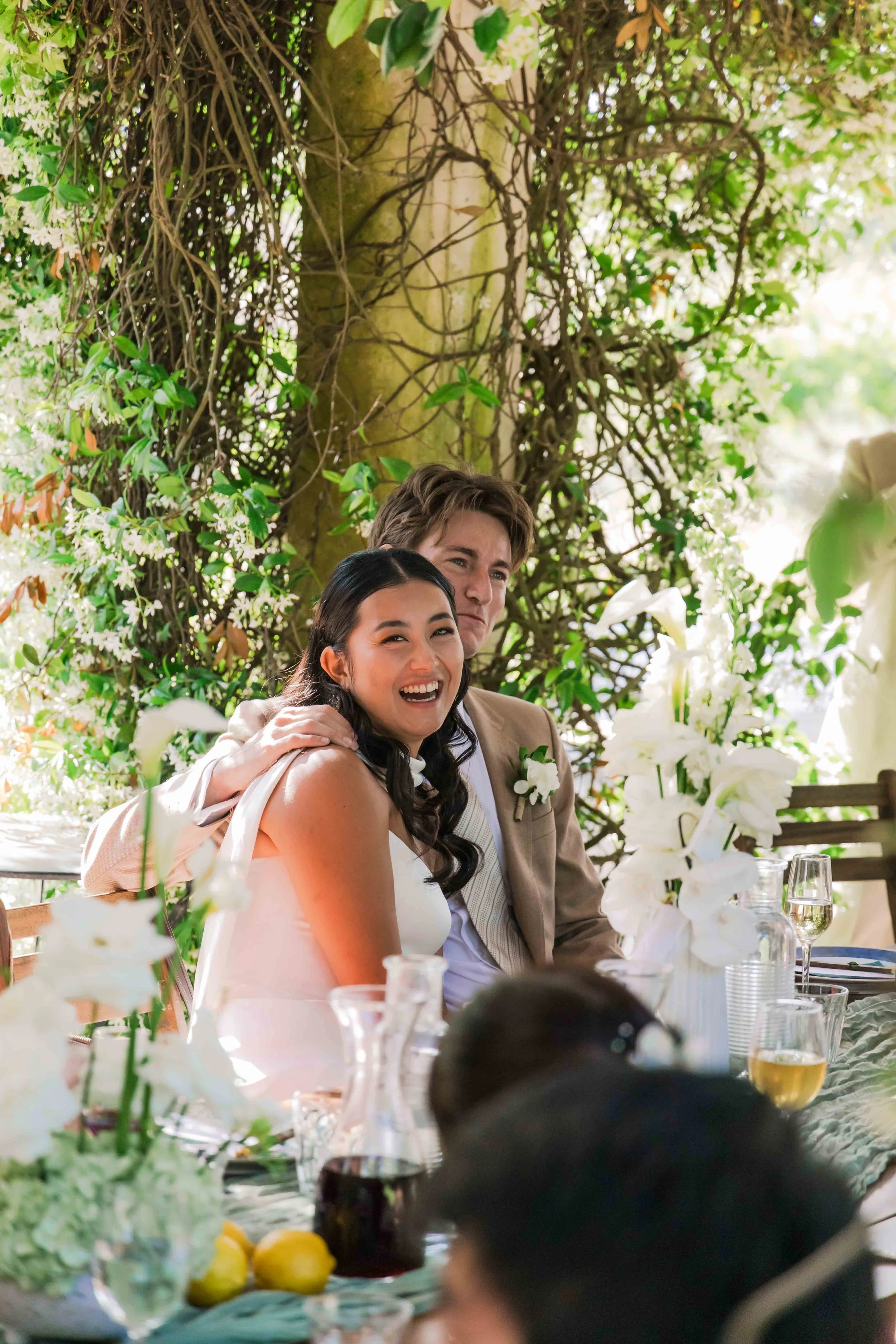 A smiling woman and a man sitting at a table outdoors during a wedding reception, surrounded by greenery and white flowers.