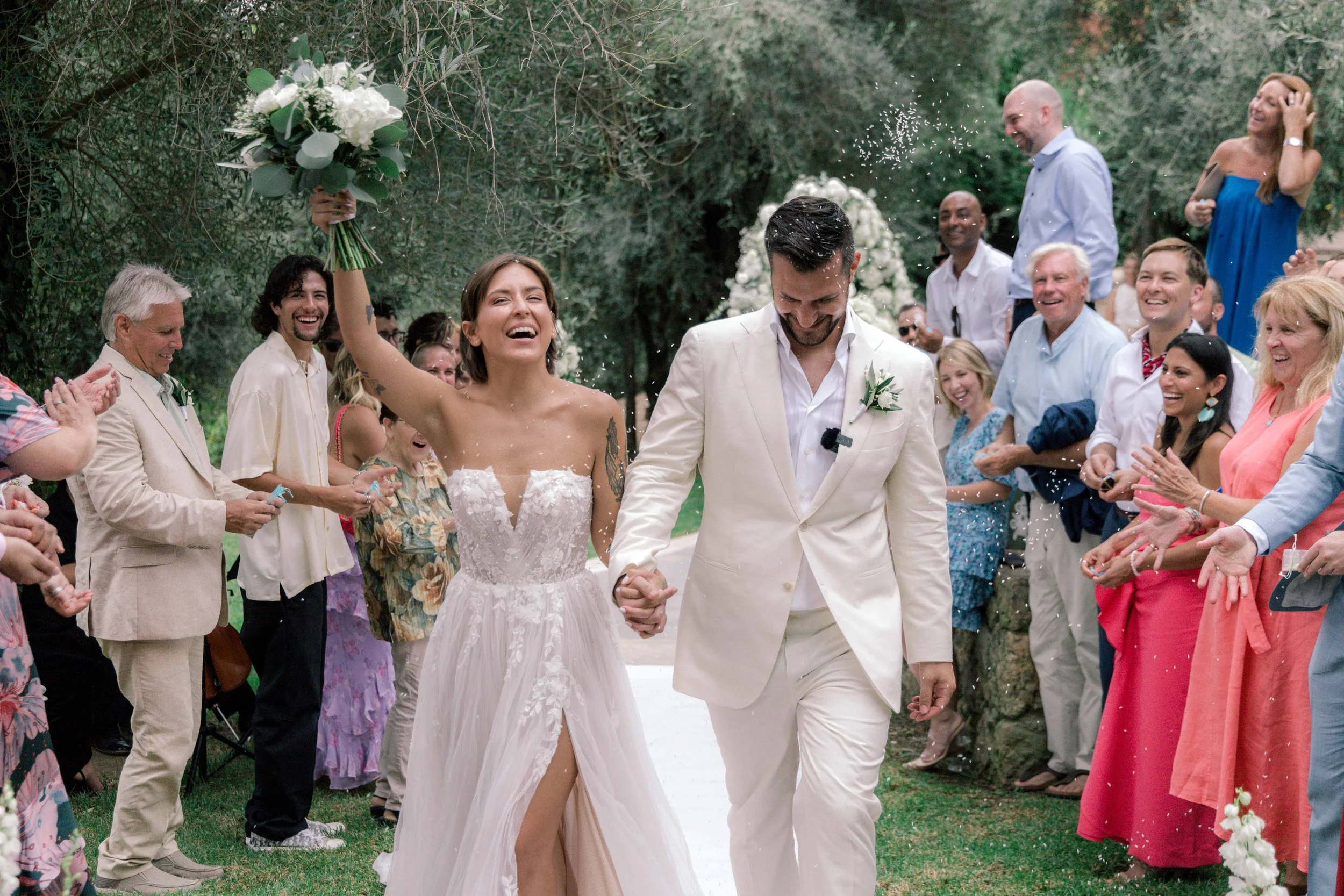 A newlywed couple walking hand in hand down an outdoor aisle, surrounded by smiling guests. The bride is in a strapless wedding dress with a slit, holding a bouquet, while the groom is in a white suit. White flower petals are being thrown, and the scene is set in a lush green garden with trees and floral arrangements.
