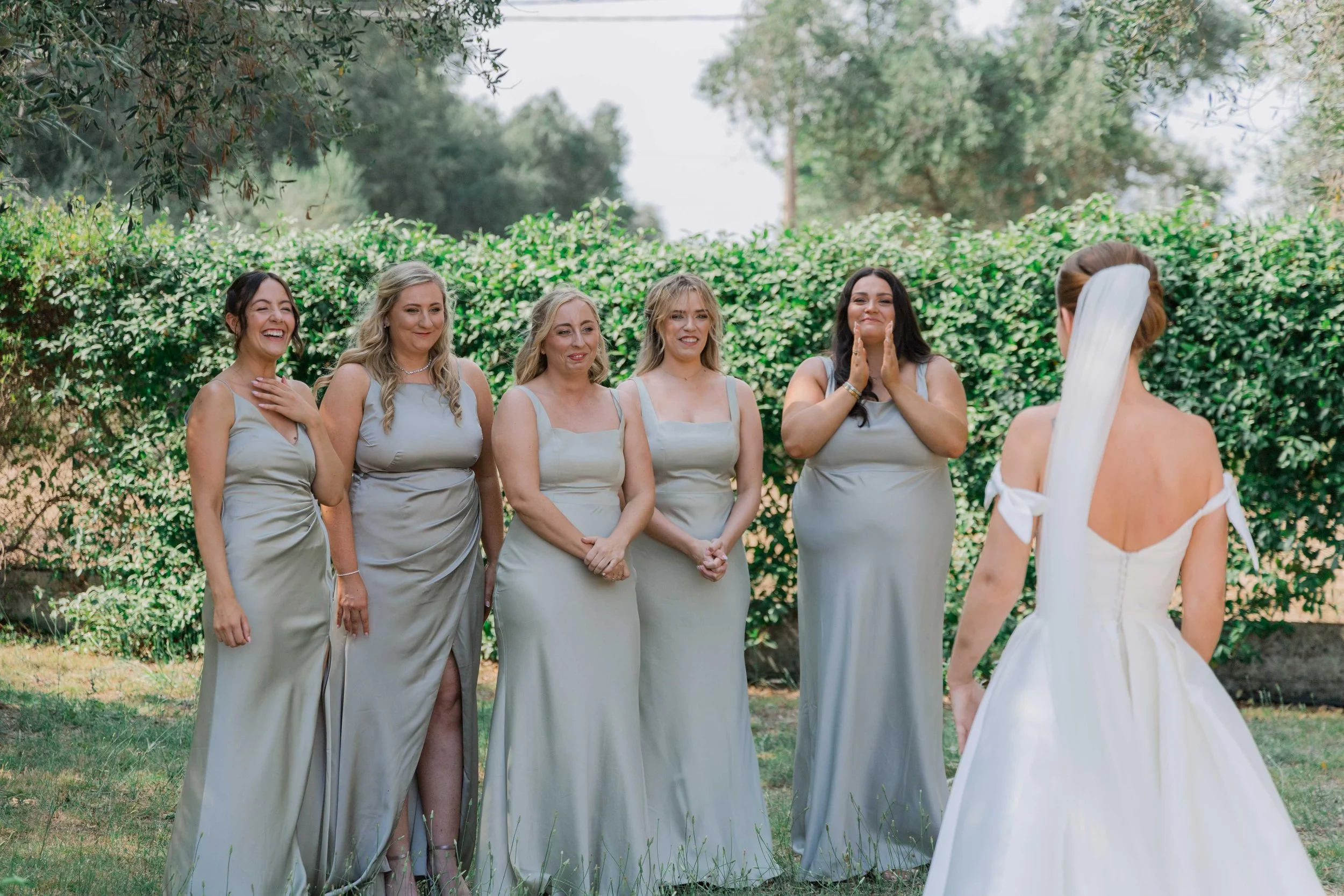 A bride in a white wedding dress with a long veil facing five bridesmaids in light gray dresses, standing outdoors in front of green shrubbery and trees, with the bridesmaids reacting emotionally to the bride.