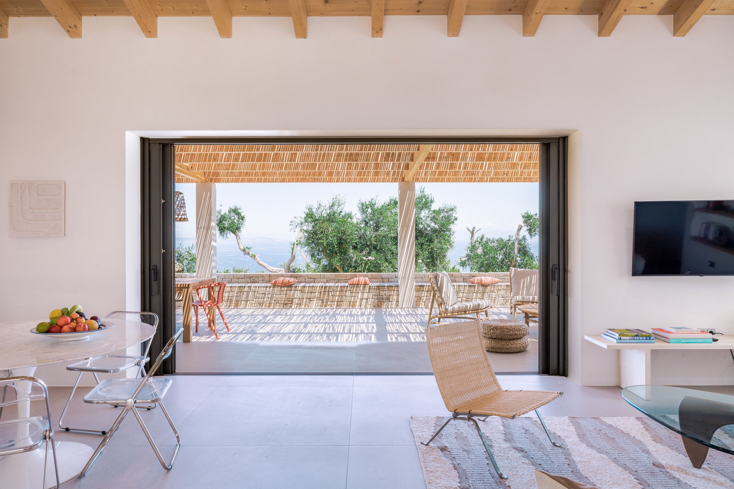 Modern living room with sliding glass door leading to balcony, outdoor chairs, trees, and view of the ocean.