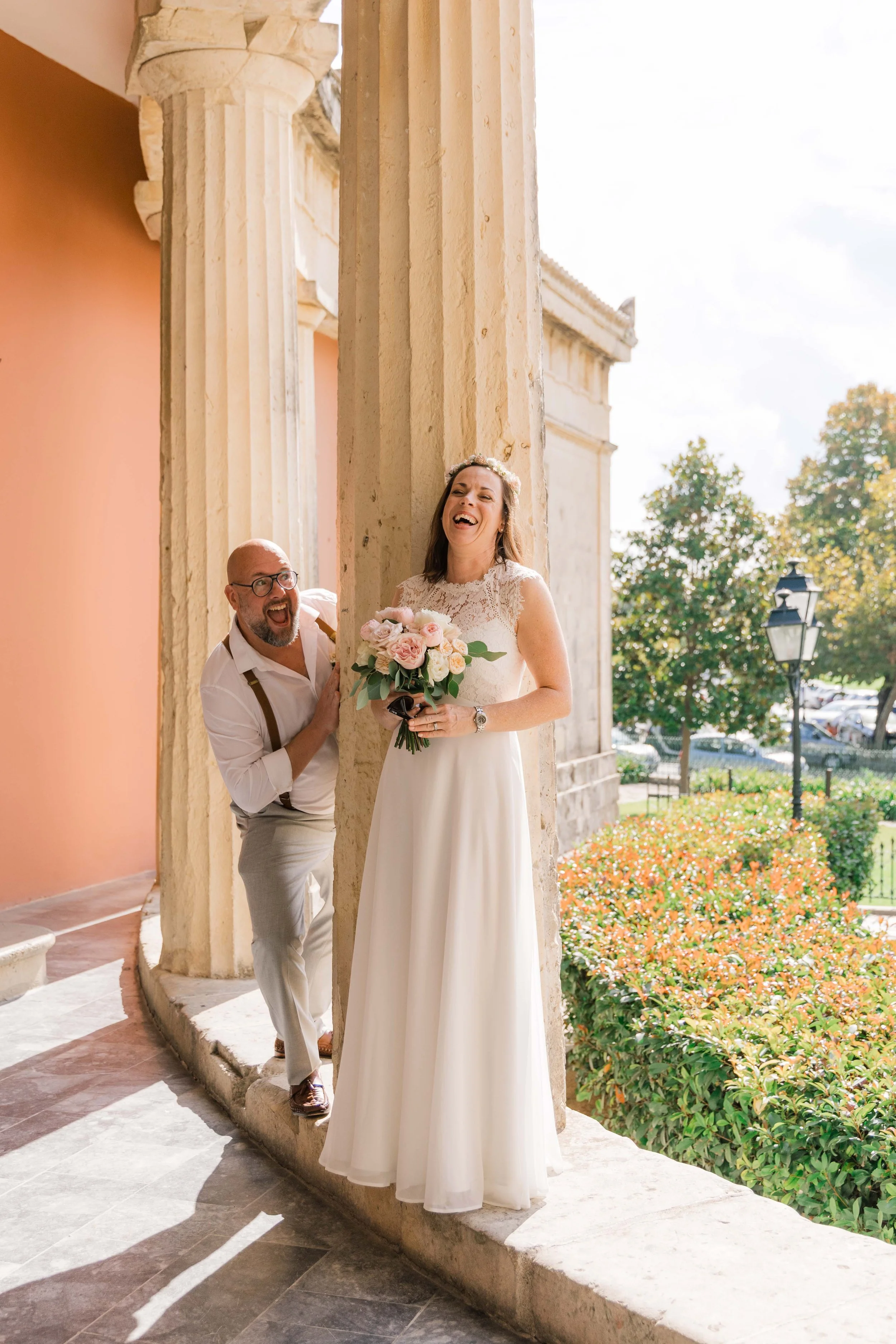 A bride in a white wedding dress holding a bouquet of pink and white flowers, laughing happily while a man peeks out from behind a stone column on her wedding day, outdoors with trees and a pathway in the background.