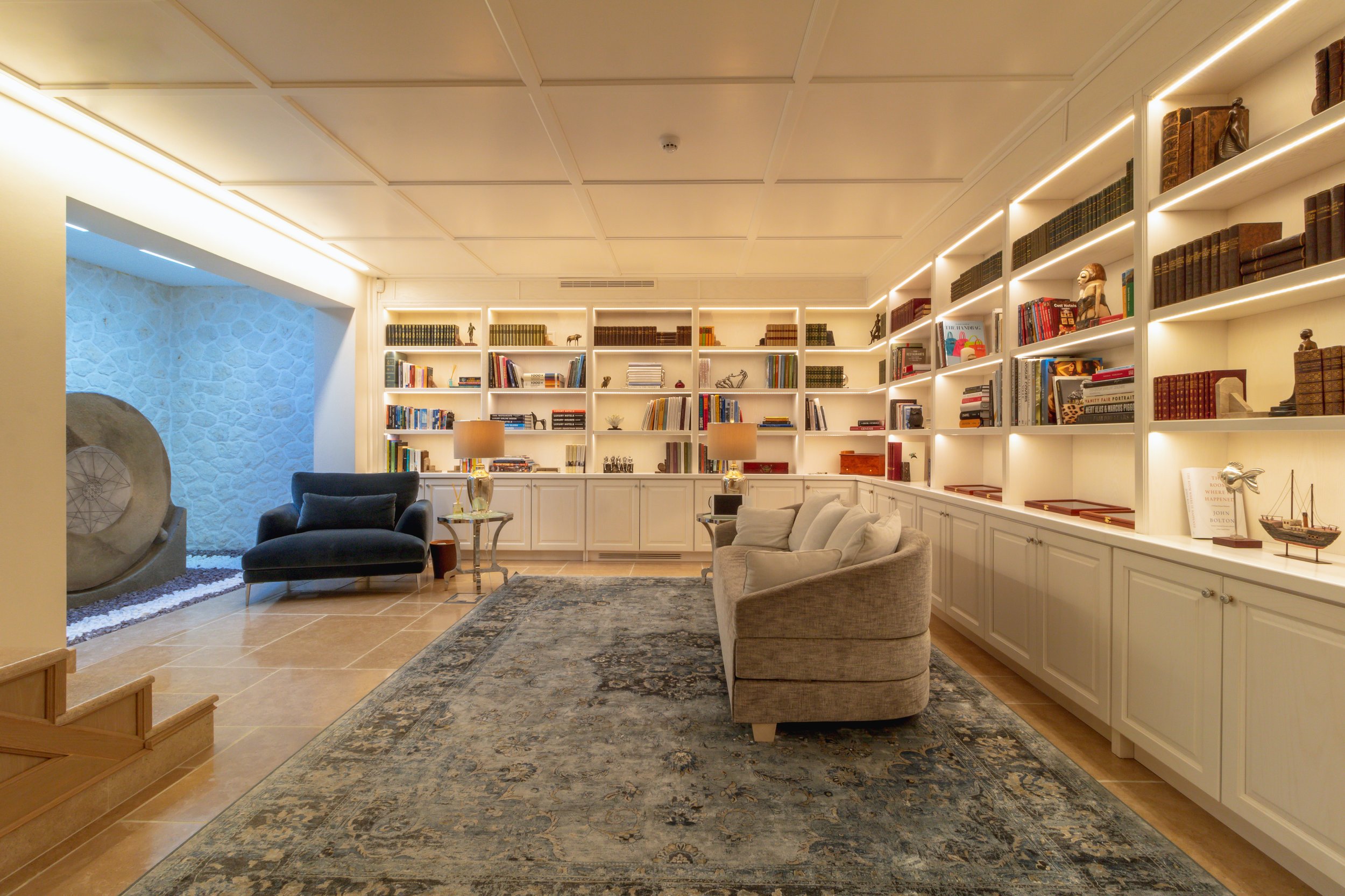 A cozy living room with white built-in shelves filled with books, a gray sofa, a dark blue armchair, and a patterned rug.