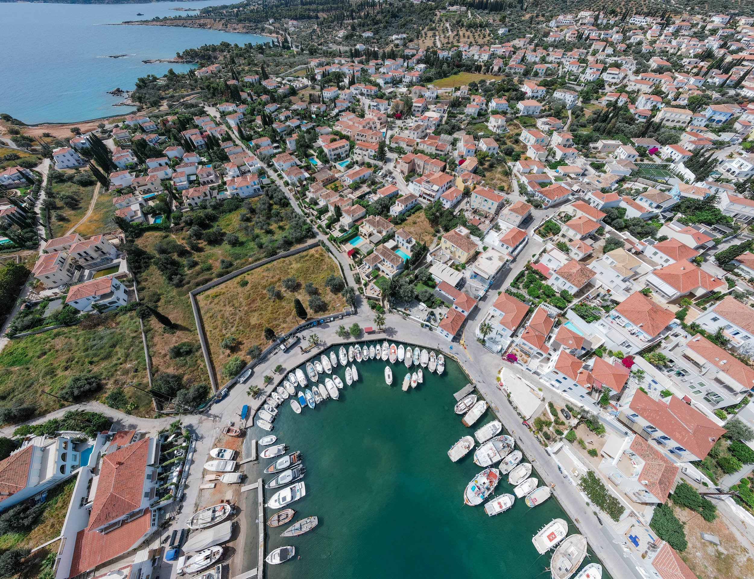 Aerial view of a coastal Mediterranean town with a small marina filled with boats, surrounded by residential buildings with red-tiled roofs, greenery, and a waterfront in the distance.