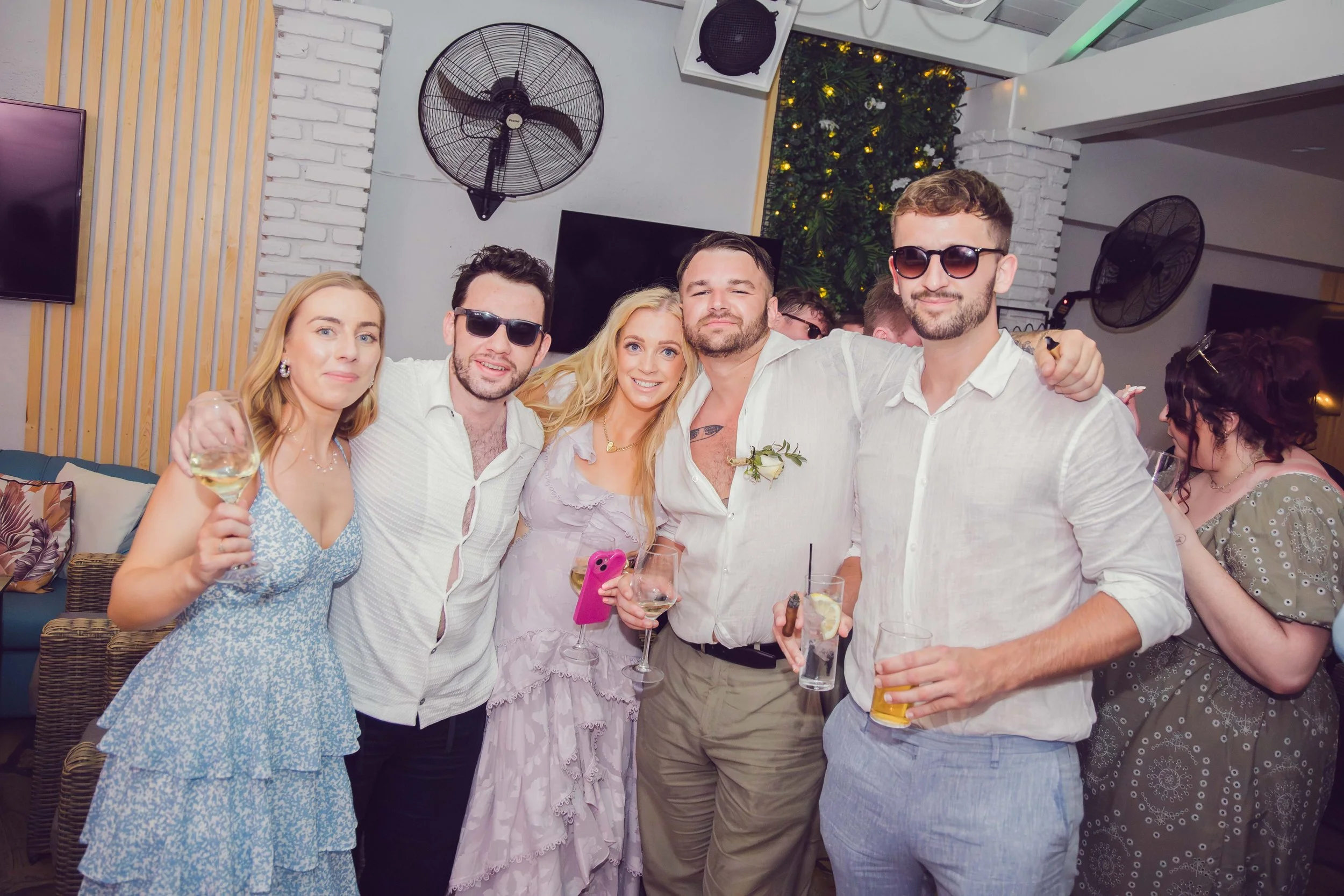 A group of six adults at a social gathering, smiling and holding drinks. The setting appears festive with decorative lighting and greenery in the background.