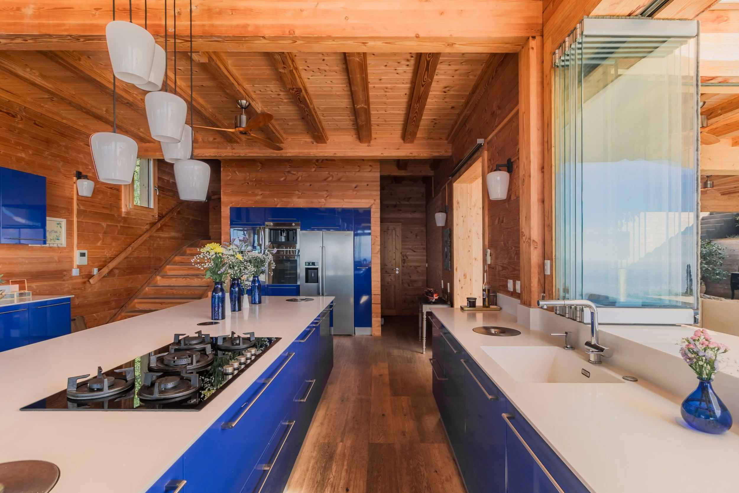 Kitchen with wooden walls and ceiling, blue cabinets, white countertops, a stovetop, stainless steel refrigerator, hanging white pendant lights, a sliding glass door, and a vase with flowers.