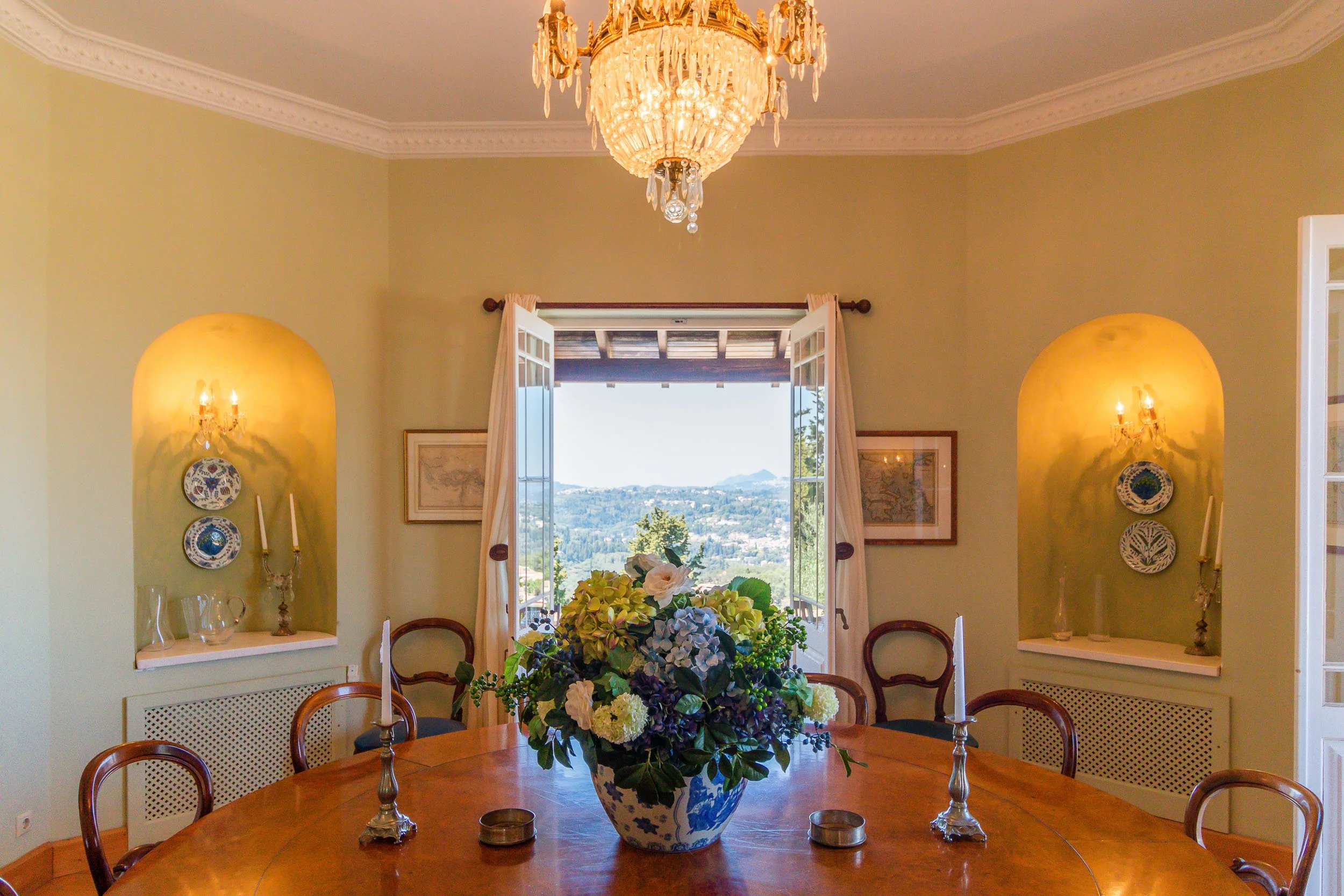 Dining room with a round wooden table, floral centerpiece in a blue and white vase, and candleholders. Open window showing scenic landscape of hills and trees in the distance. Elegant chandelier hanging from ceiling, two wall niches with decorative p