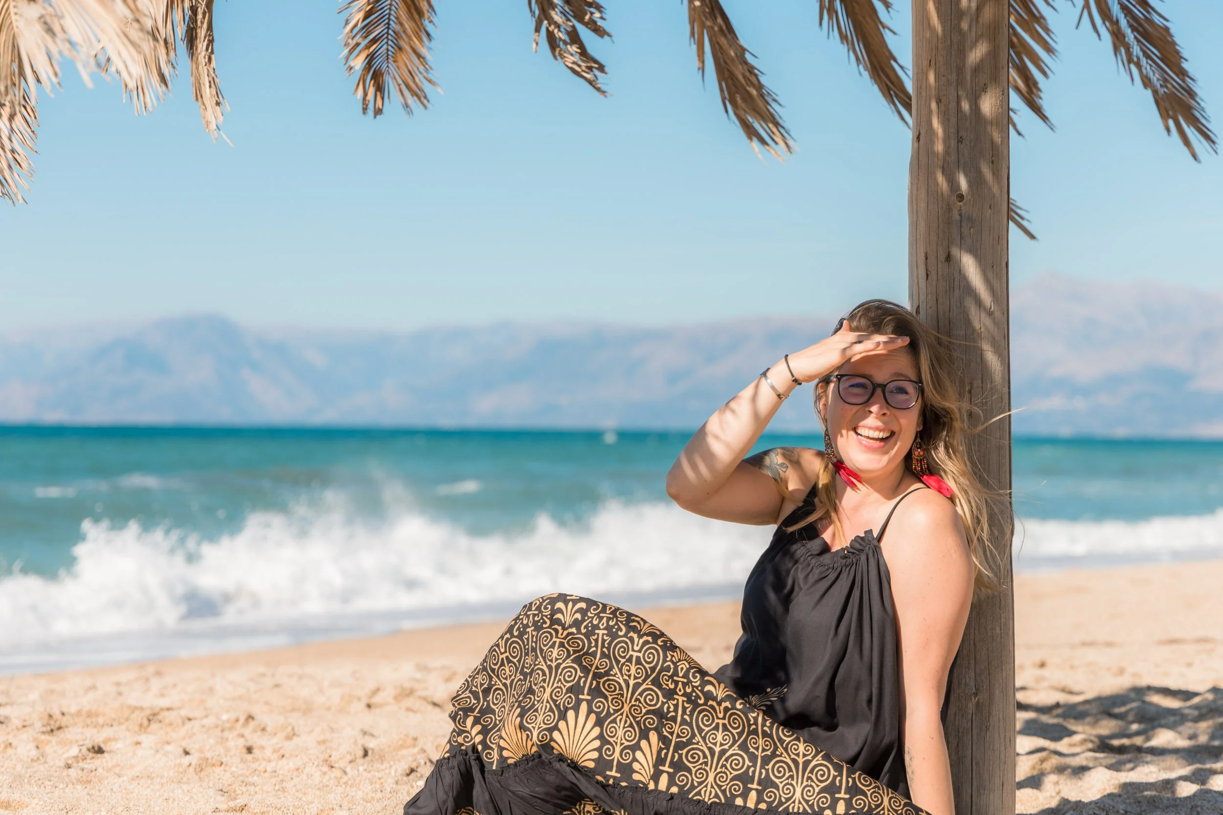 A smiling woman in sunglasses and a black dress sitting on a beach under a thatched umbrella, shielding her eyes from the sun with her hand, with ocean waves and mountains in the background.