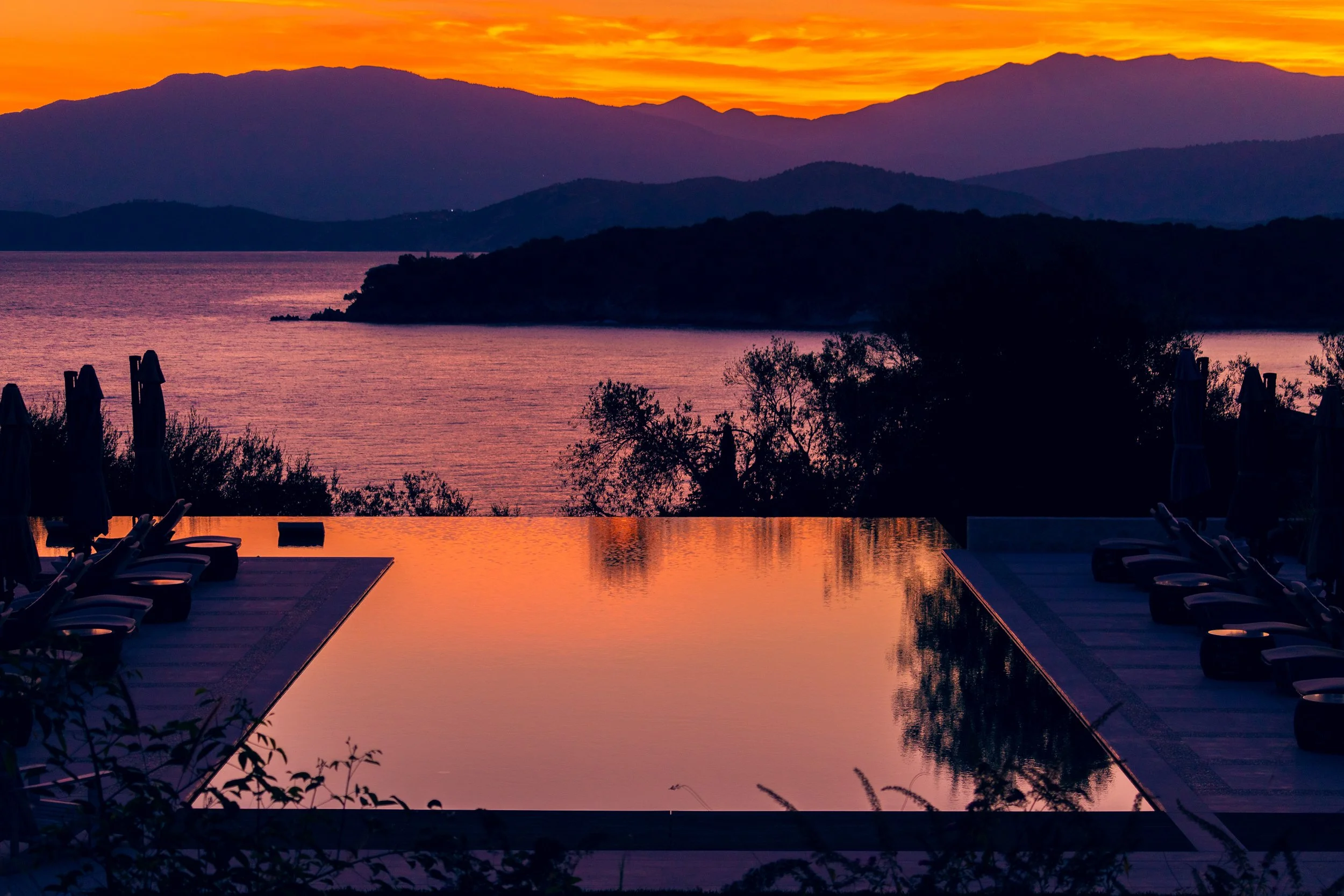 Sunset over mountains and lake, with an infinity pool and lounge chairs in the foreground.