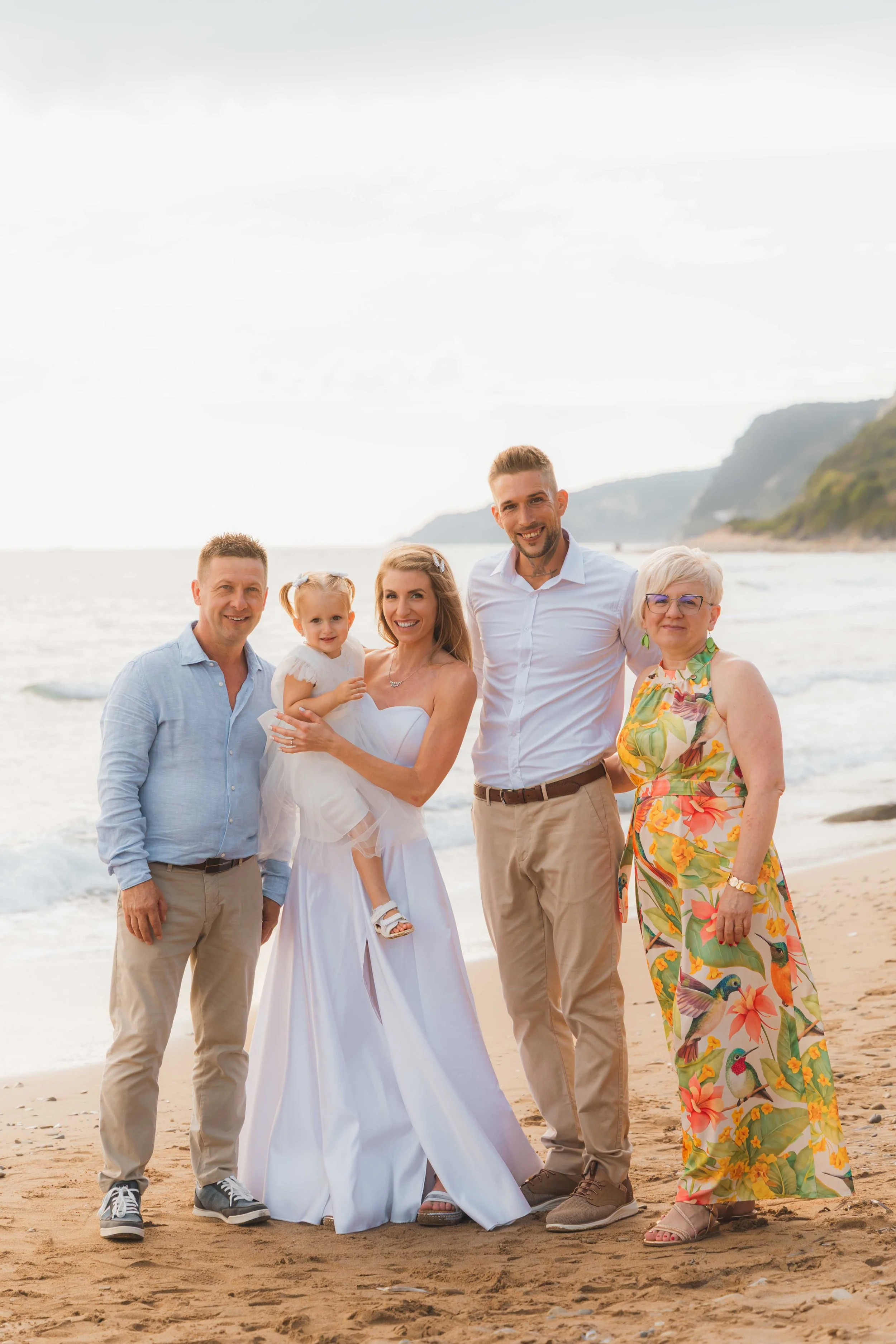 A family at the beach during sunset, with five people, including a young girl in a white dress being held by a woman in a white gown, all smiling.