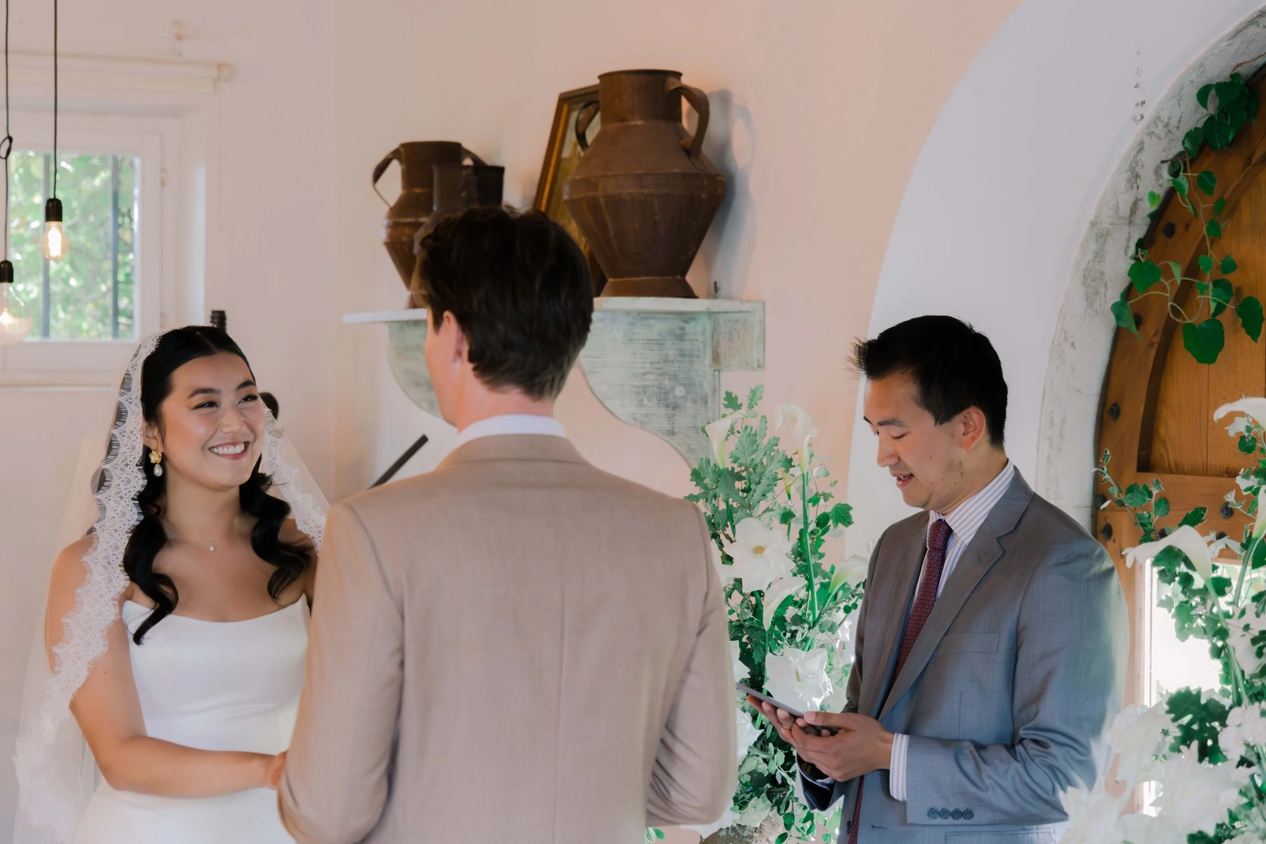 Bride and groom exchanging vows during their wedding ceremony, with an officiant reading from a tablet, in a decorated indoor venue with flowers and antique vases.