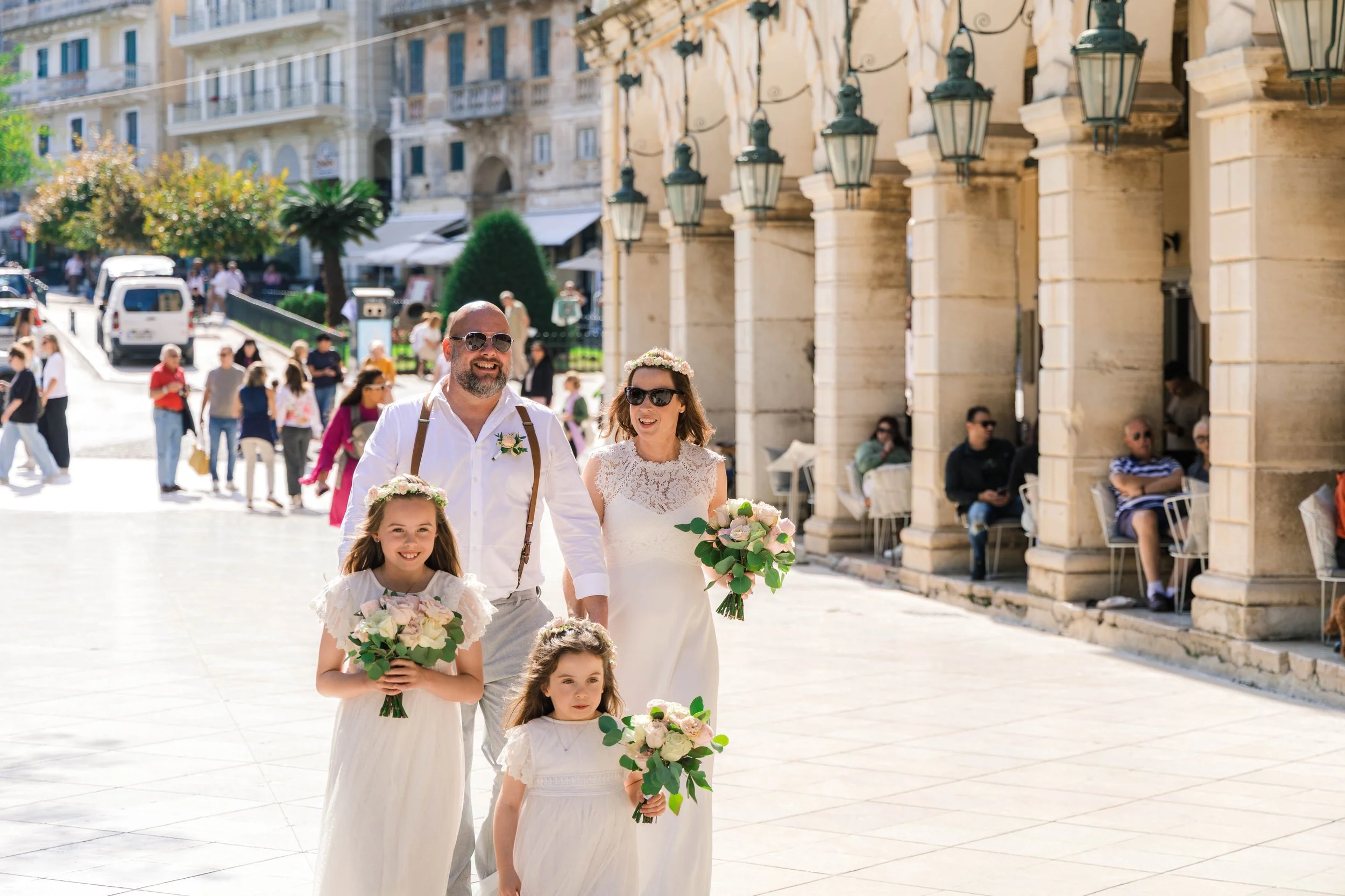 A family celebrating a wedding outdoors on a sunny day, with a bride and groom and two young girls holding bouquets of flowers, walking on a city street with people and historic buildings in the background.