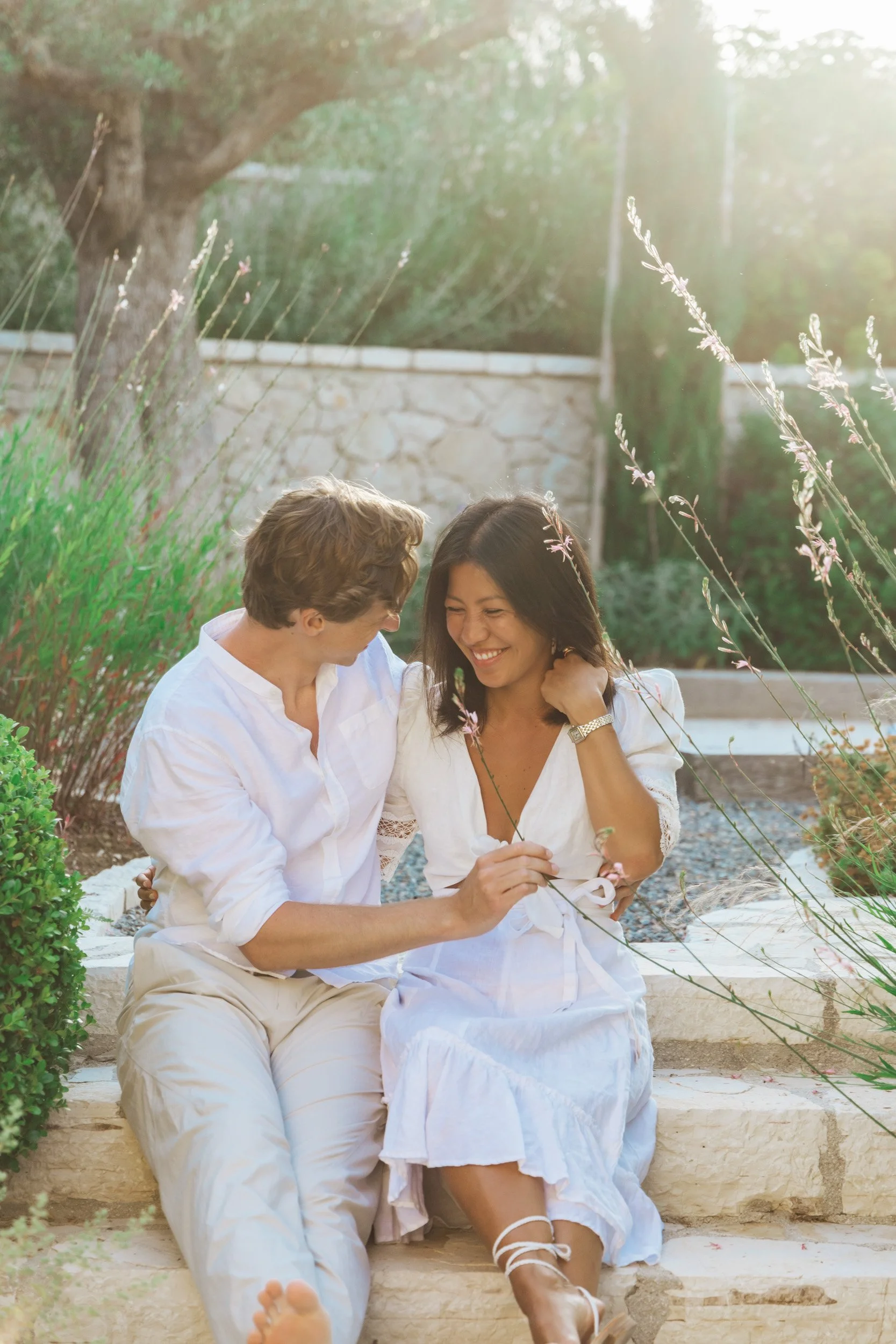 A smiling couple sitting outdoors on stone steps, surrounded by plants, during sunset, with the man holding a flower for the woman.