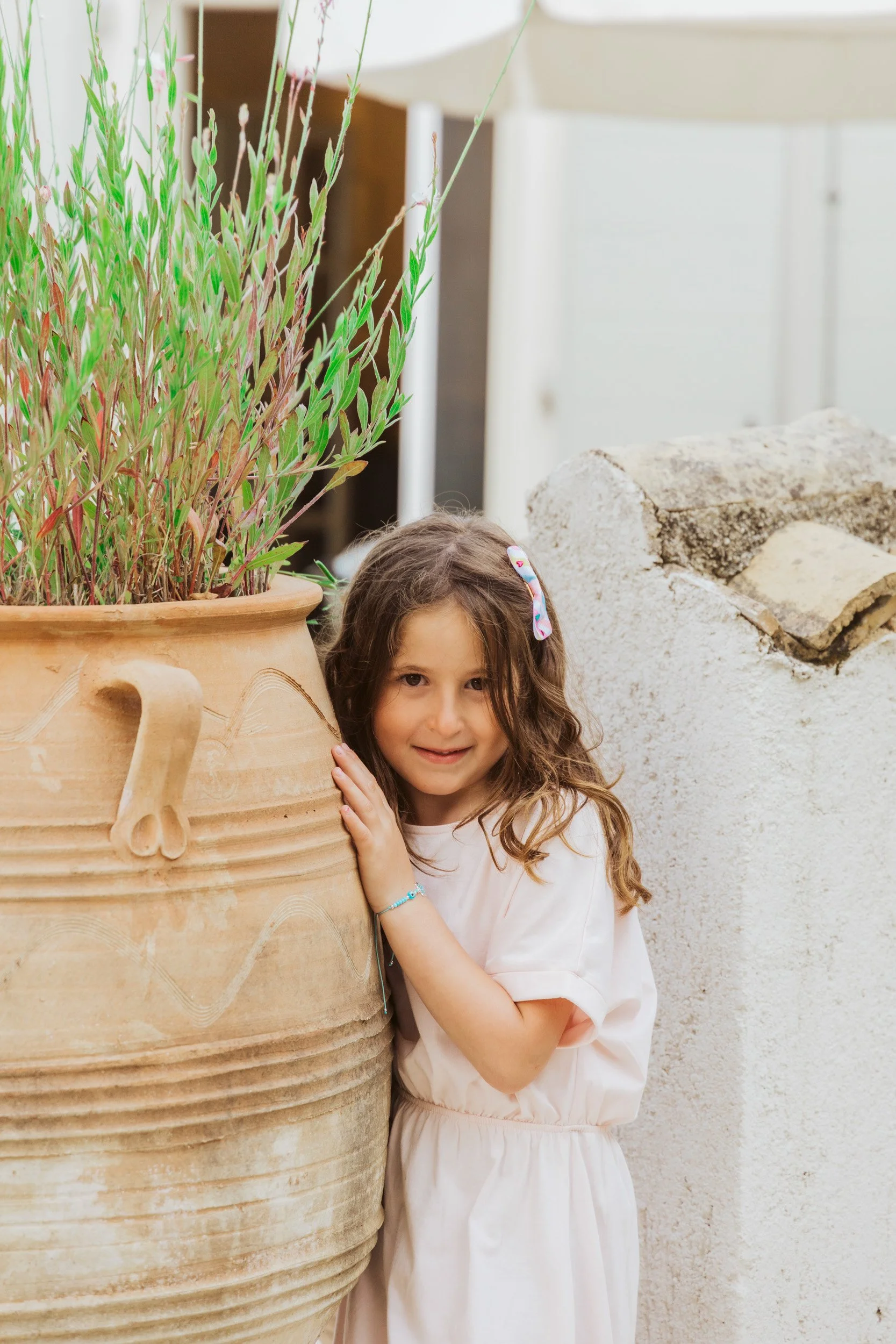 A young girl with long brown hair, wearing a light pink dress and a colorful hair clip, is hugging a large terracotta pot with green plants.