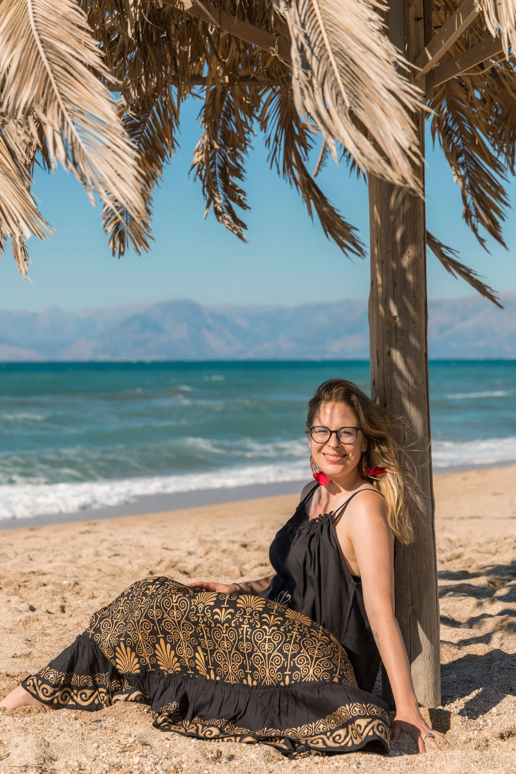 A woman with glasses and earrings, smiling, sitting on sandy beach under a thatched umbrella, with ocean and mountains in the background.