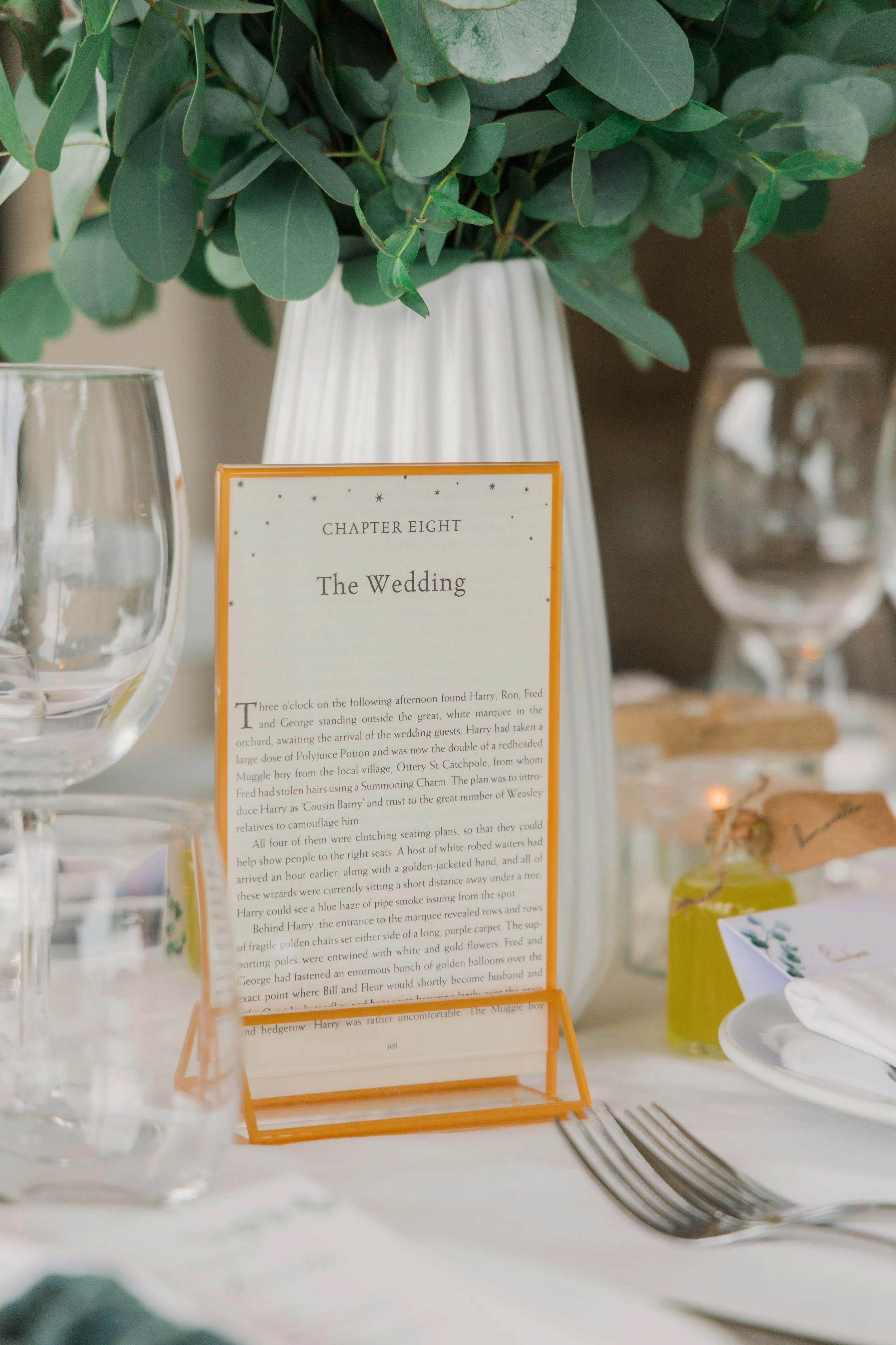 Wedding reception table with a menu card titled 'Chapter Eight: The Wedding', decorative greenery in a white vase, wine glasses, and a plate with a folded napkin and small bottles of flavored oils or dressings.