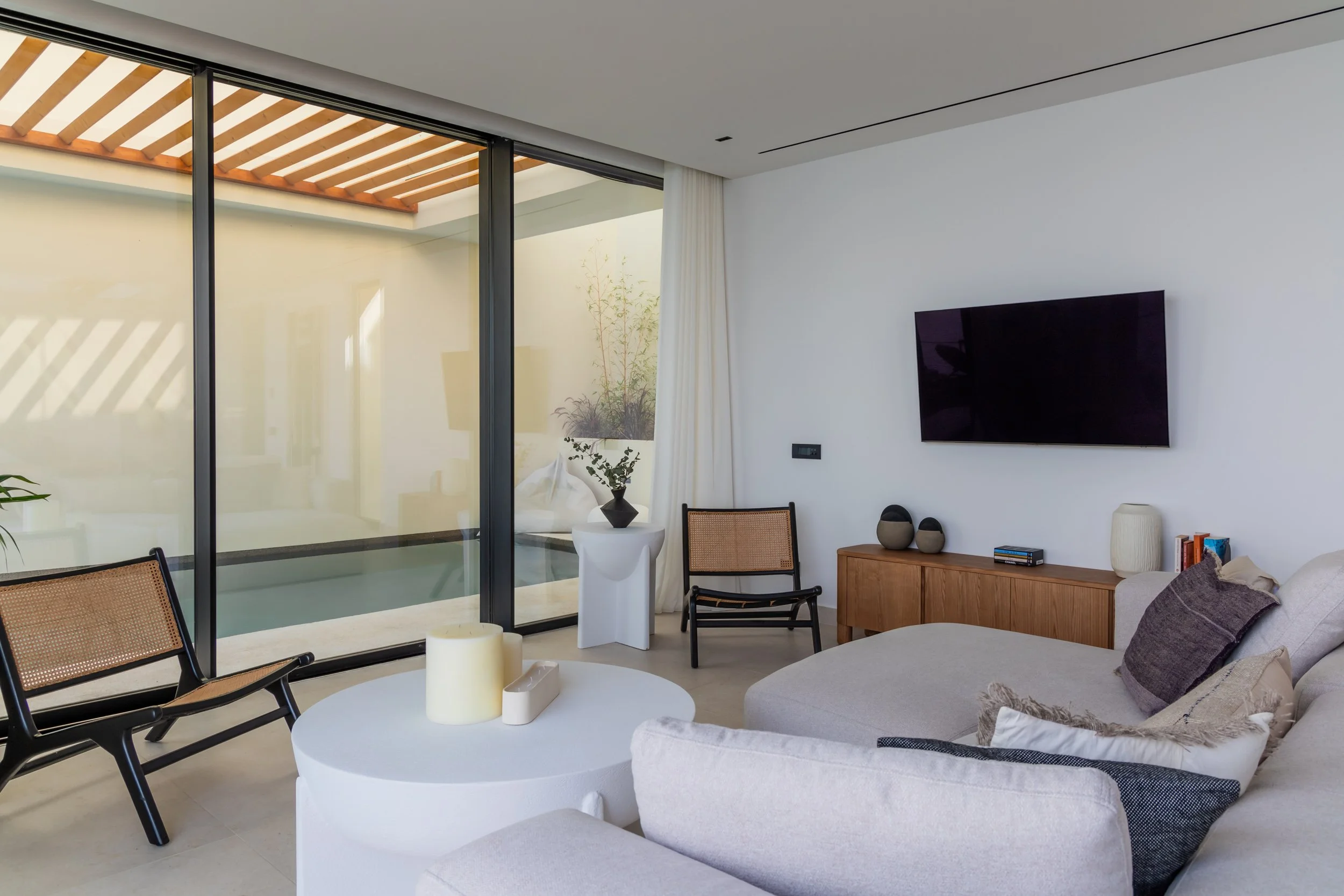 Modern living room with a beige sectional sofa, a white round coffee table with candles, black armchairs with woven cane backrests, a wooden media console, wall-mounted flat-screen TV, large glass sliding doors leading outside, and decor including va