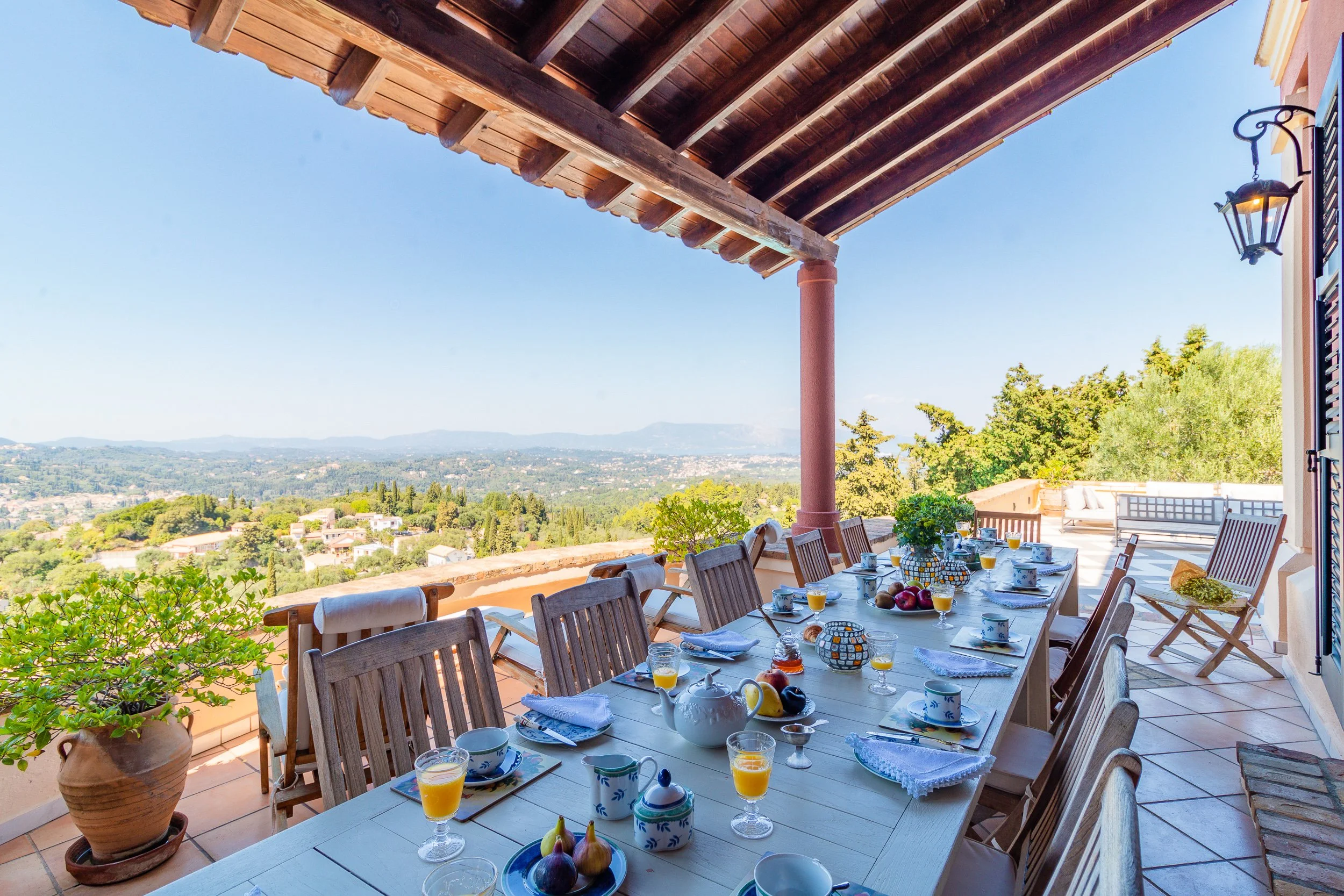 Outdoor dining table set on a terrace overlooking a scenic green landscape and distant hills, with a partly covered wooden pergola overhead.