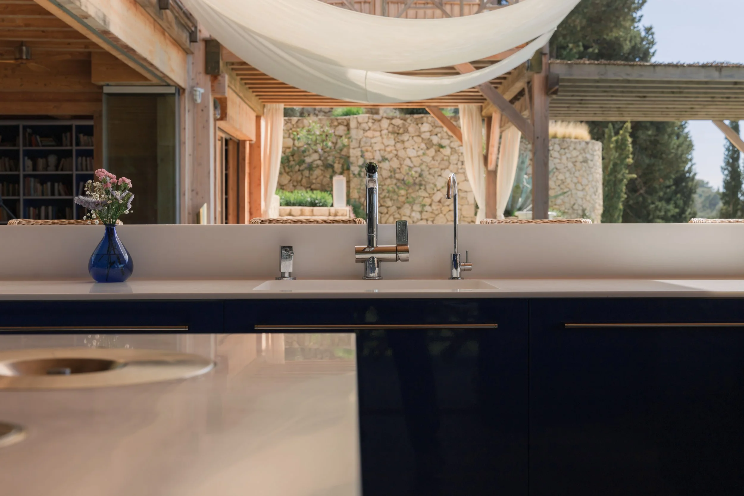 Kitchen sink with a blue vase containing pink and white flowers, set in front of a window showing an outdoor patio with stone wall, trees, and wooden pergola with hanging fabric.