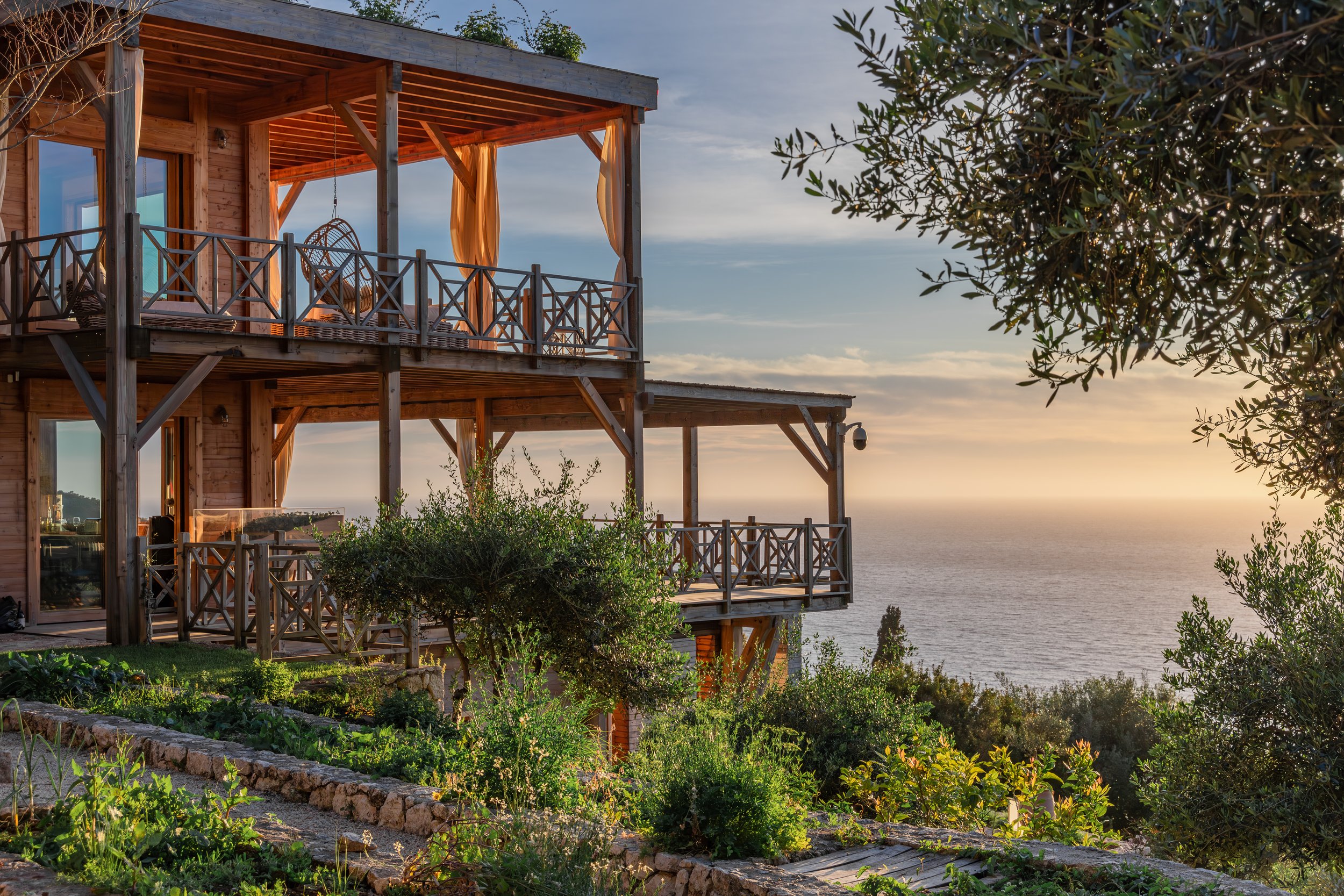 A wooden multi-level house with an outdoor balcony overlooking the ocean during sunset, surrounded by green plants and trees.