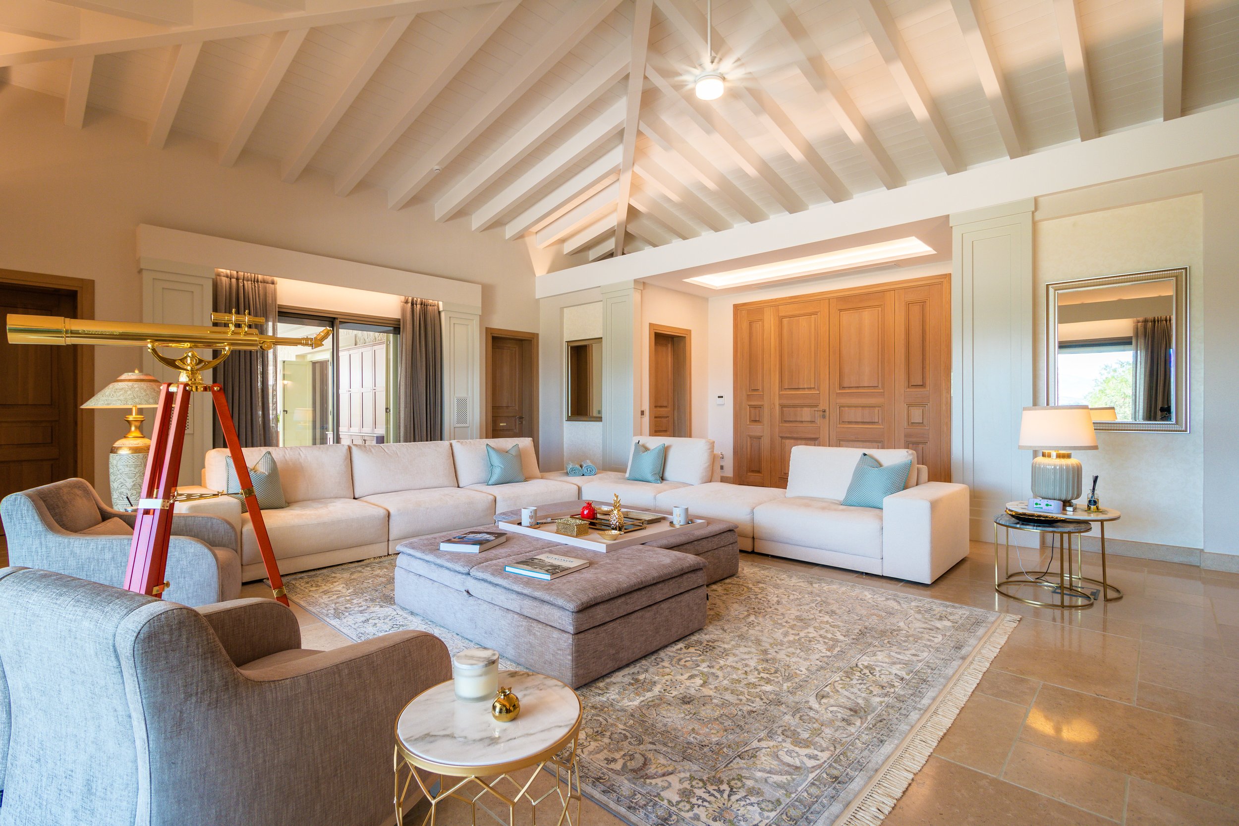 A spacious living room with high vaulted white ceiling, large white sectional sofa with blue pillows, a beige armchair, a marble-topped side table with a candle, a brass lamp, and a large central ottoman with books and decorations, decorated in neutr