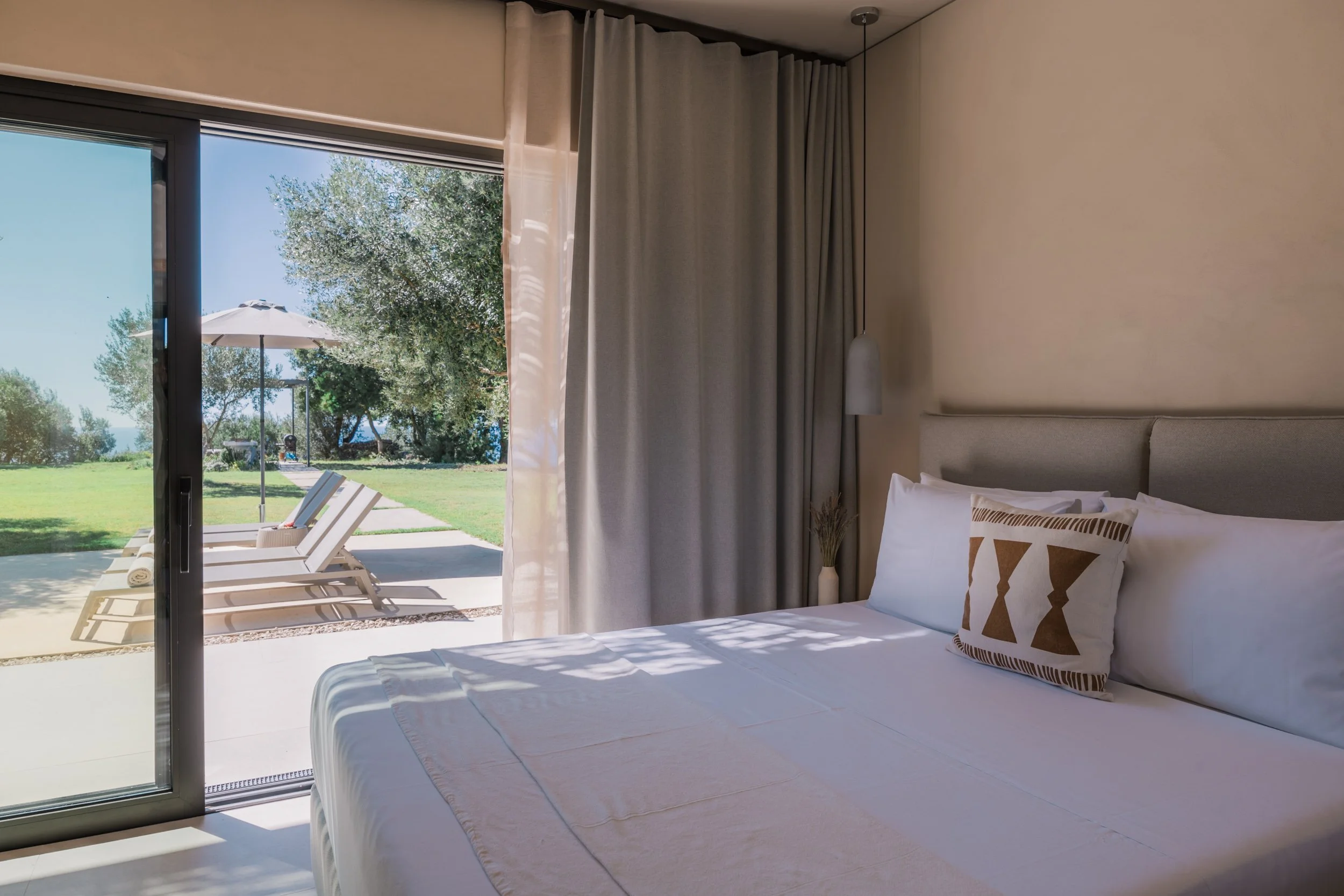 A bedroom with a large bed, light-colored bedding, decorative pillow, hanging pendant light, and a sliding glass door leading to an outdoor patio with lounge chairs and a sun umbrella, overlooking a grassy area with trees.