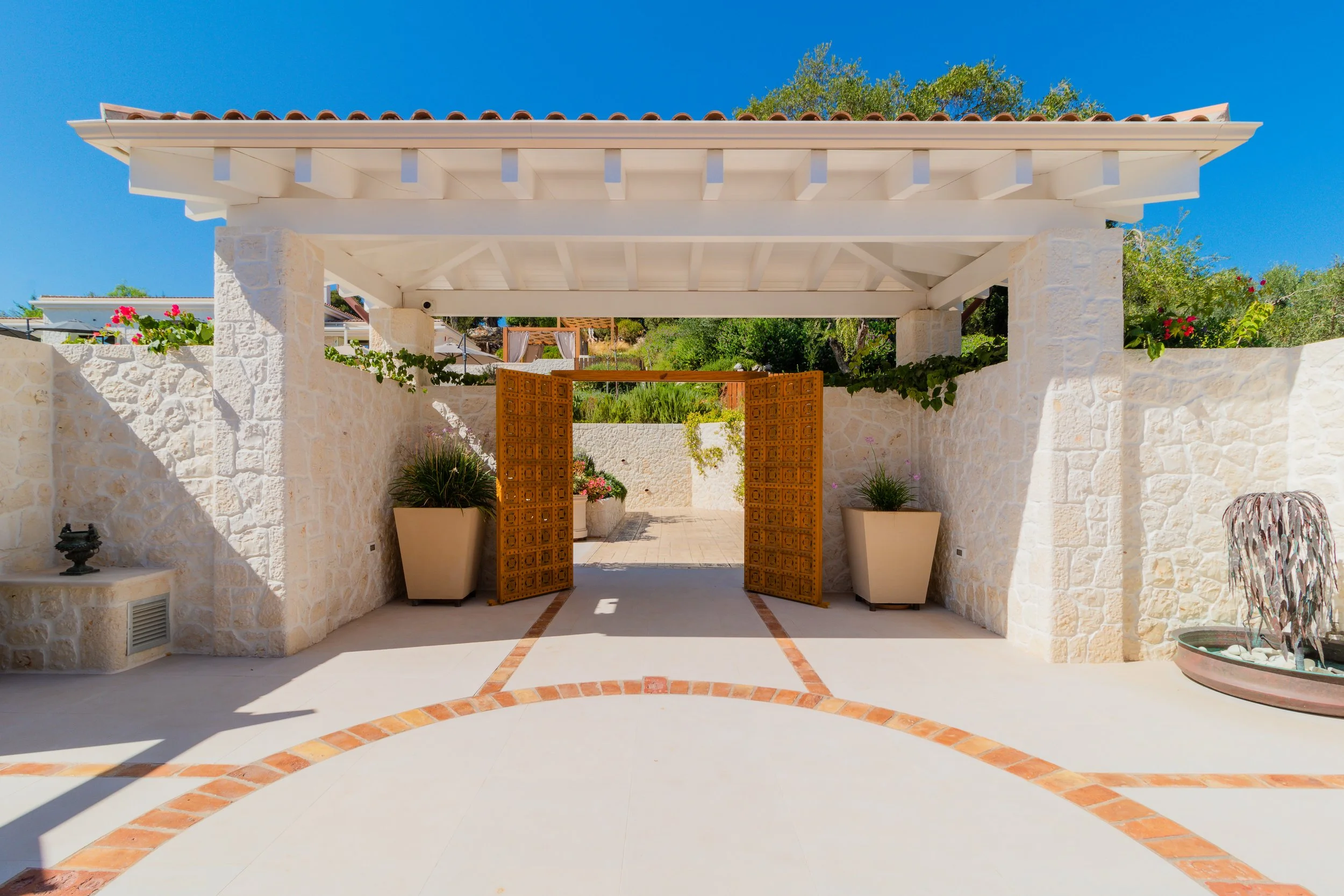 A covered outdoor patio with white stone walls, a tiled roof, and decorative tile accents on the ground leading to a wooden gate, surrounded by potted plants and greenery.