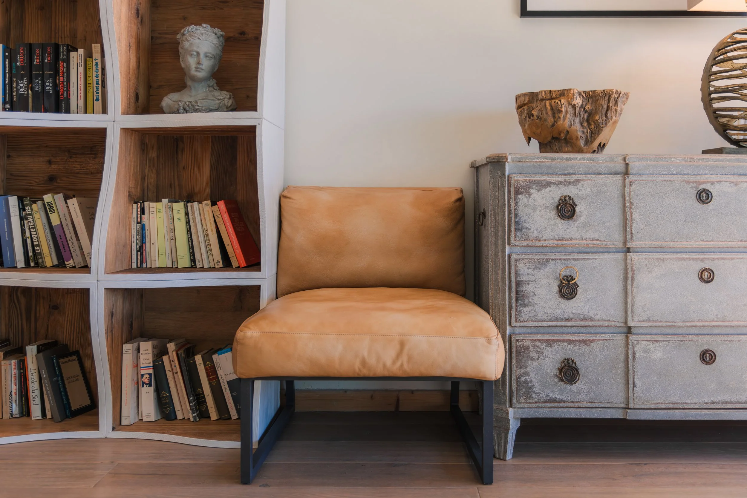 Interior scene with a wooden bookshelf filled with books, a beige leather chair, and a rustic grey dresser with decorative objects on top, including a wooden bowl and a sculpture bust.
