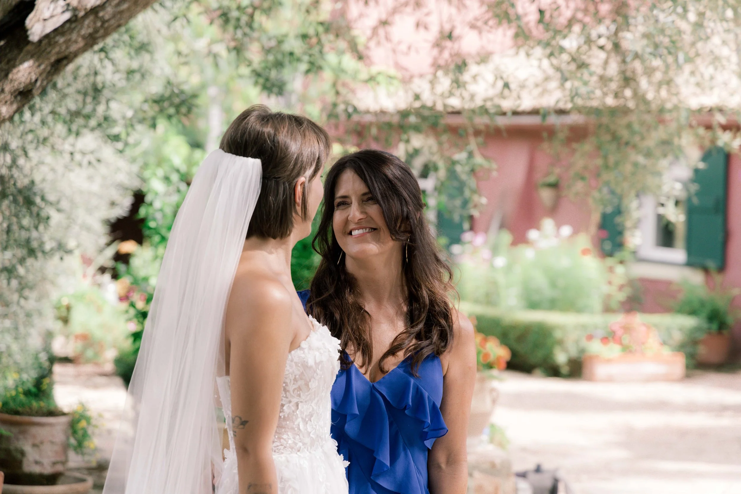 A bride in a white wedding dress with a sheer veil smiling at a woman in a blue dress outdoors under a tree with blooming flowers.