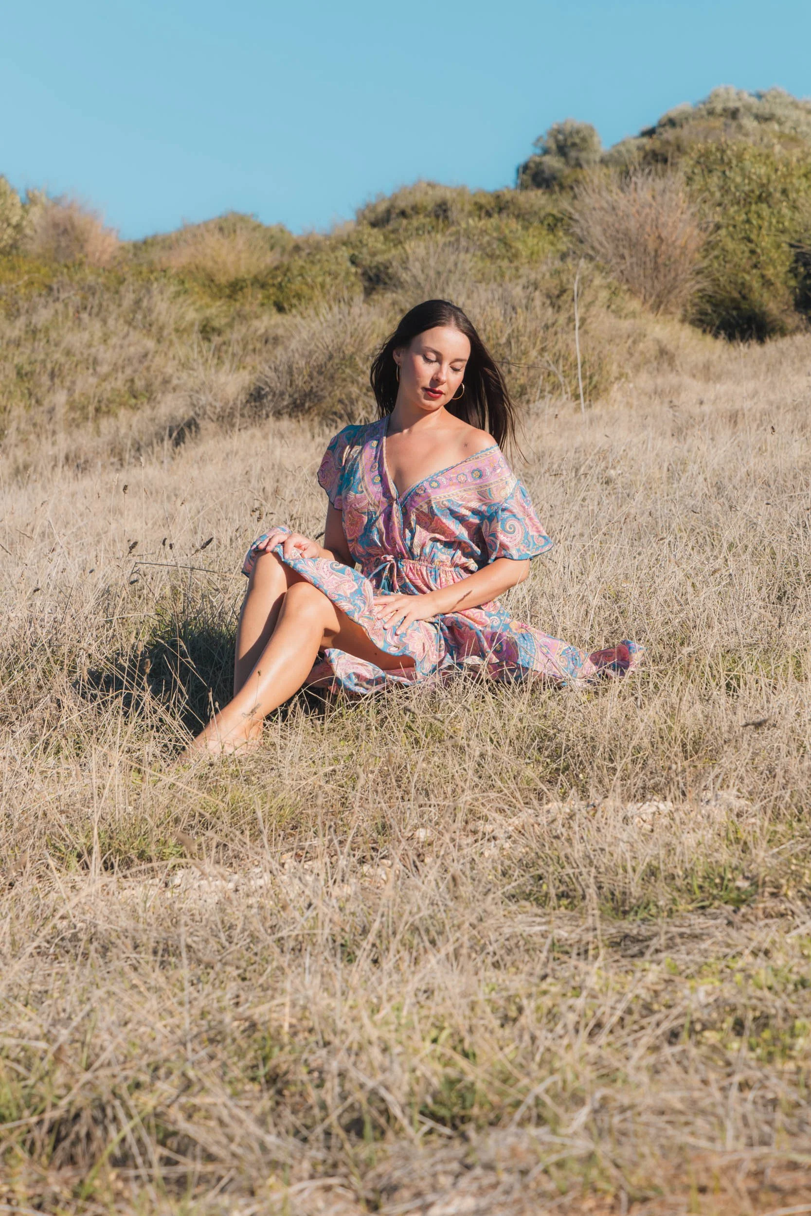 A woman sitting in a grassy field with hills and trees in the background, wearing a colorful dress and looking down.