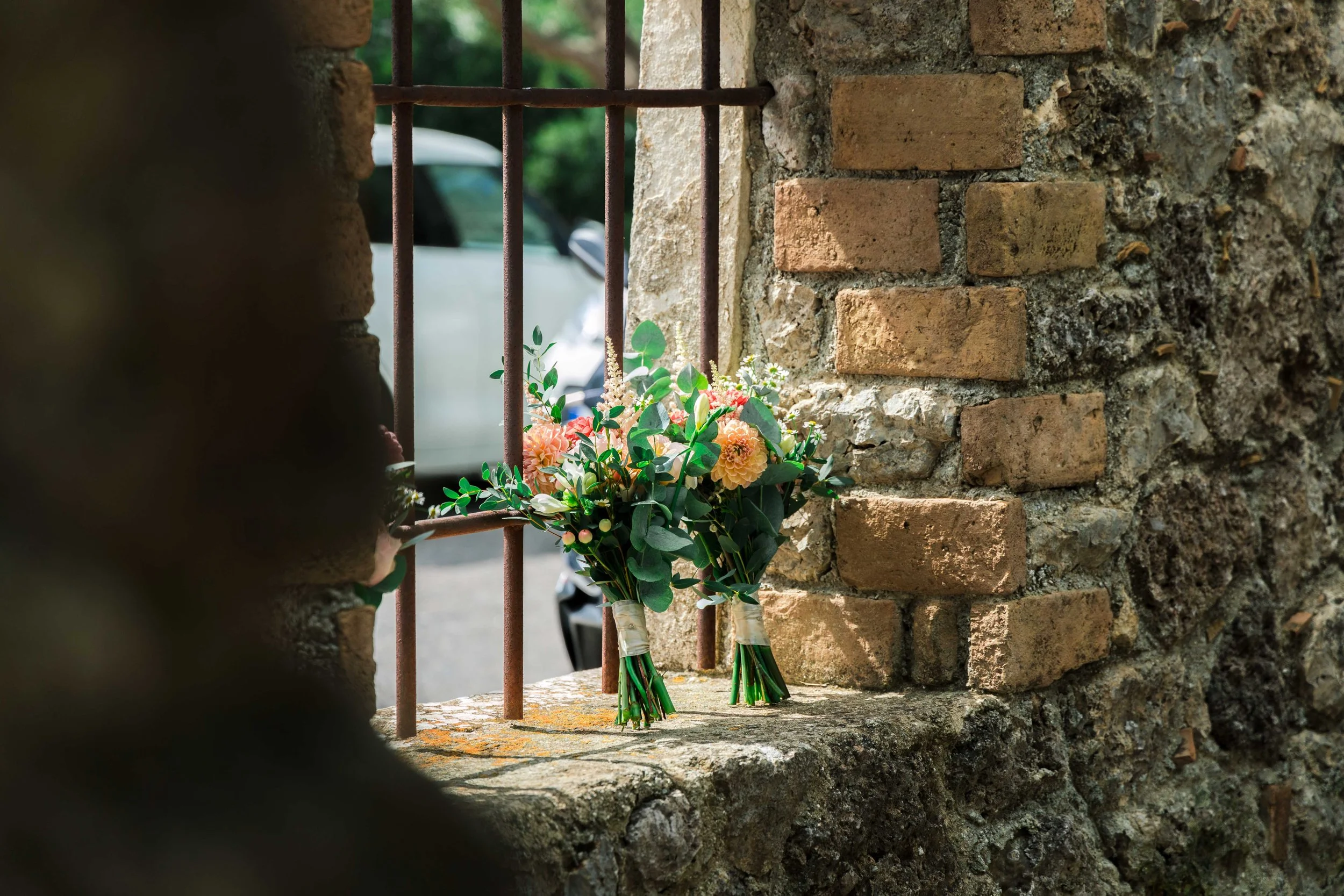 Two bouquets of peach and pink flowers tied with white ribbon, placed on a stone windowsill outside a brick and stone wall, with barred window and nearby vehicle in the background.