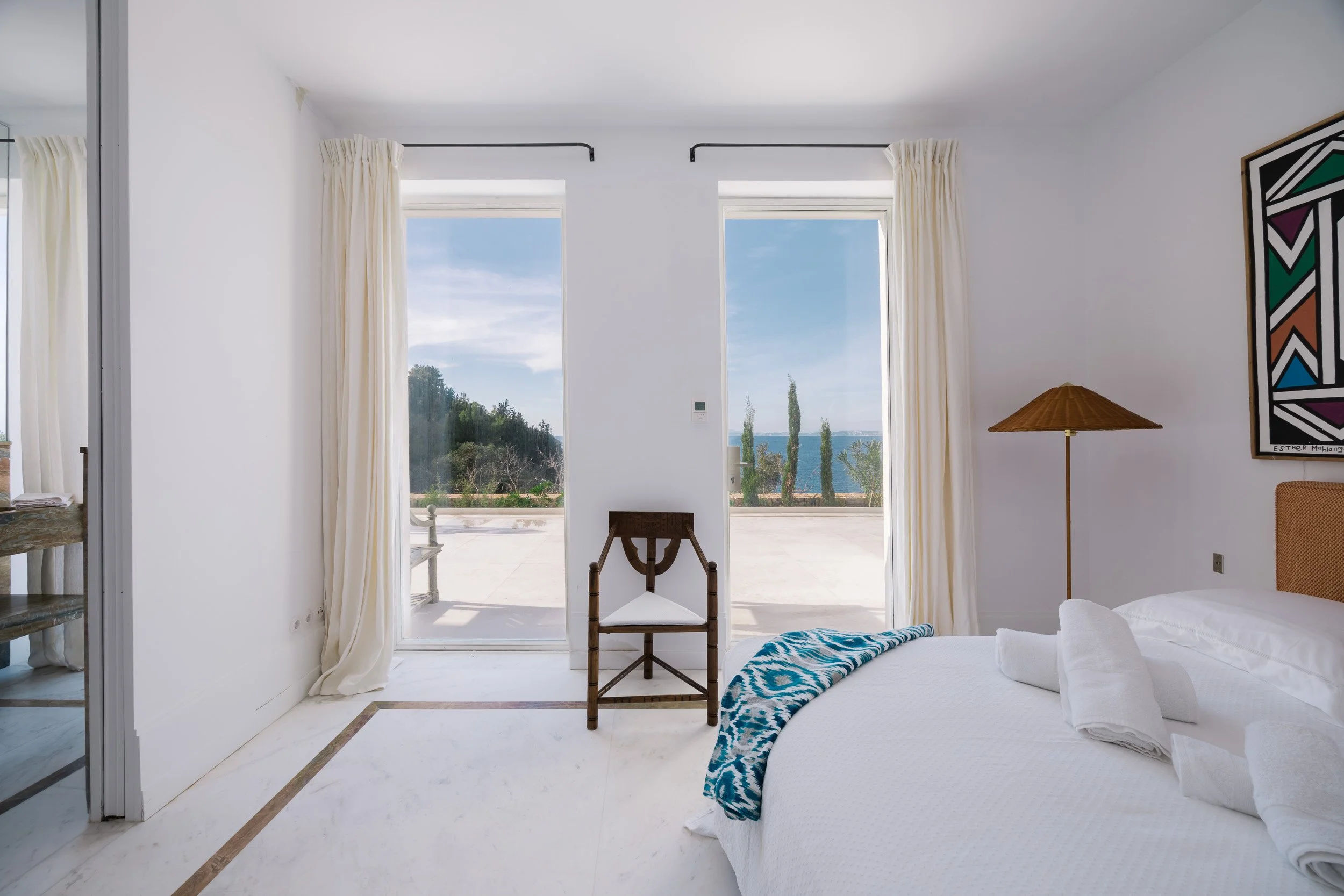 A bedroom with white walls, a bed with white linens and towels, a bedside lamp, a chair by sliding glass doors, and a view of trees, water, and the sky outside.