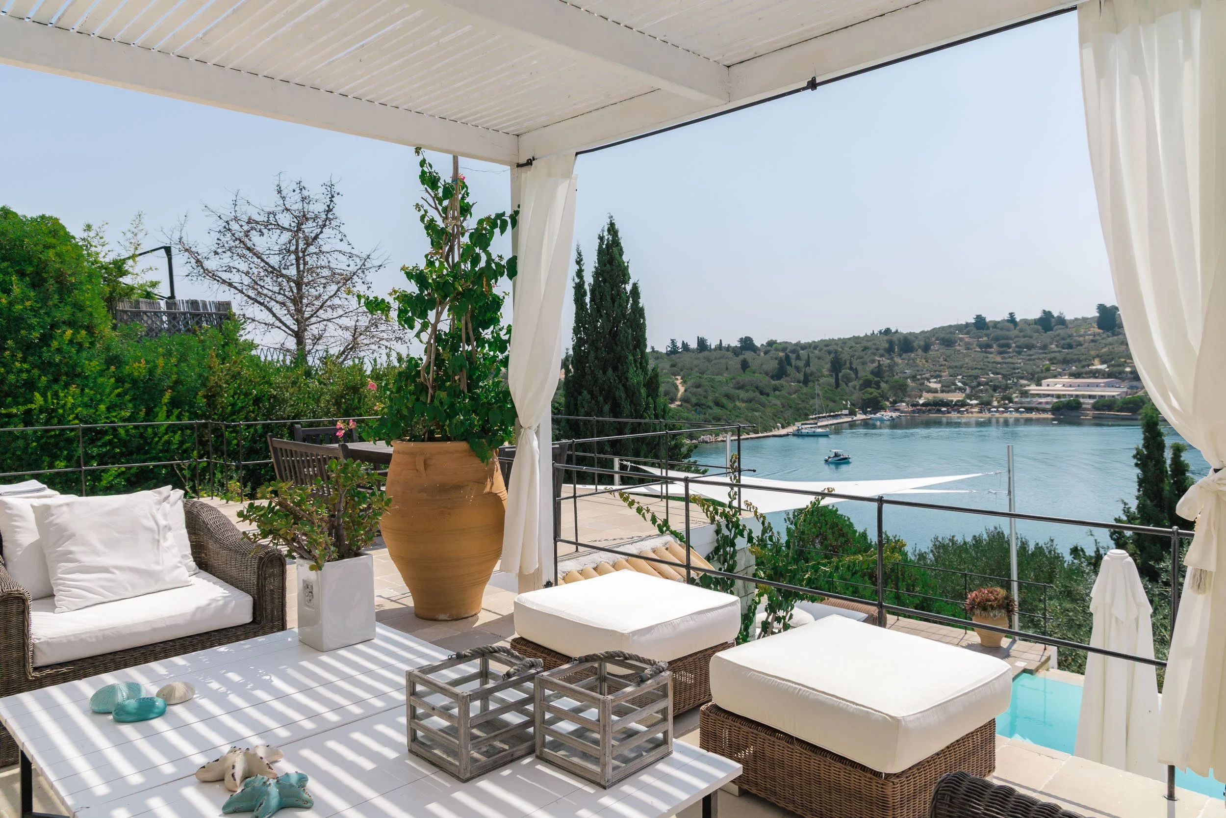 Outdoor patio with white cushions, potted plants, and a view of a calm bay with boats and a hillside in the background.