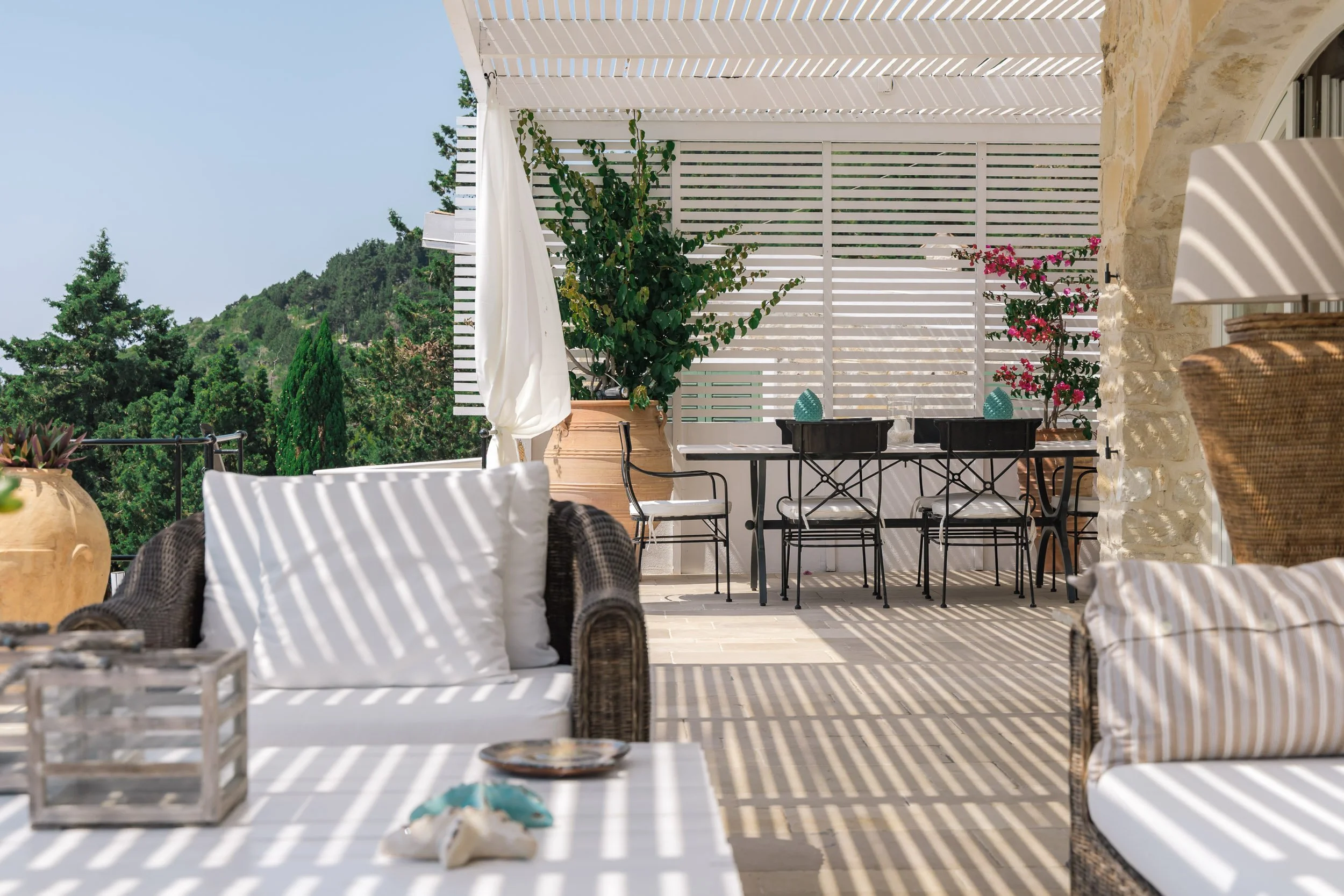Outdoor patio with wicker armchair and striped cushions, white dining table with black chairs, potted plants, and a scenic view of green trees and hills outside.