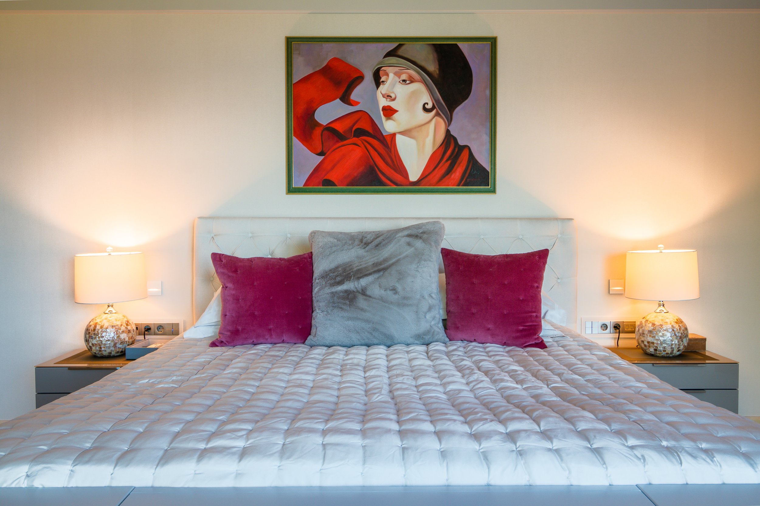 A neatly made bed with a white quilt, three pillows (two red velvet and one larger gray plush), on a white headboard. Two matching lamps with decorative bases are on bedside tables on either side of the bed. Above, a framed portrait painting of a wom