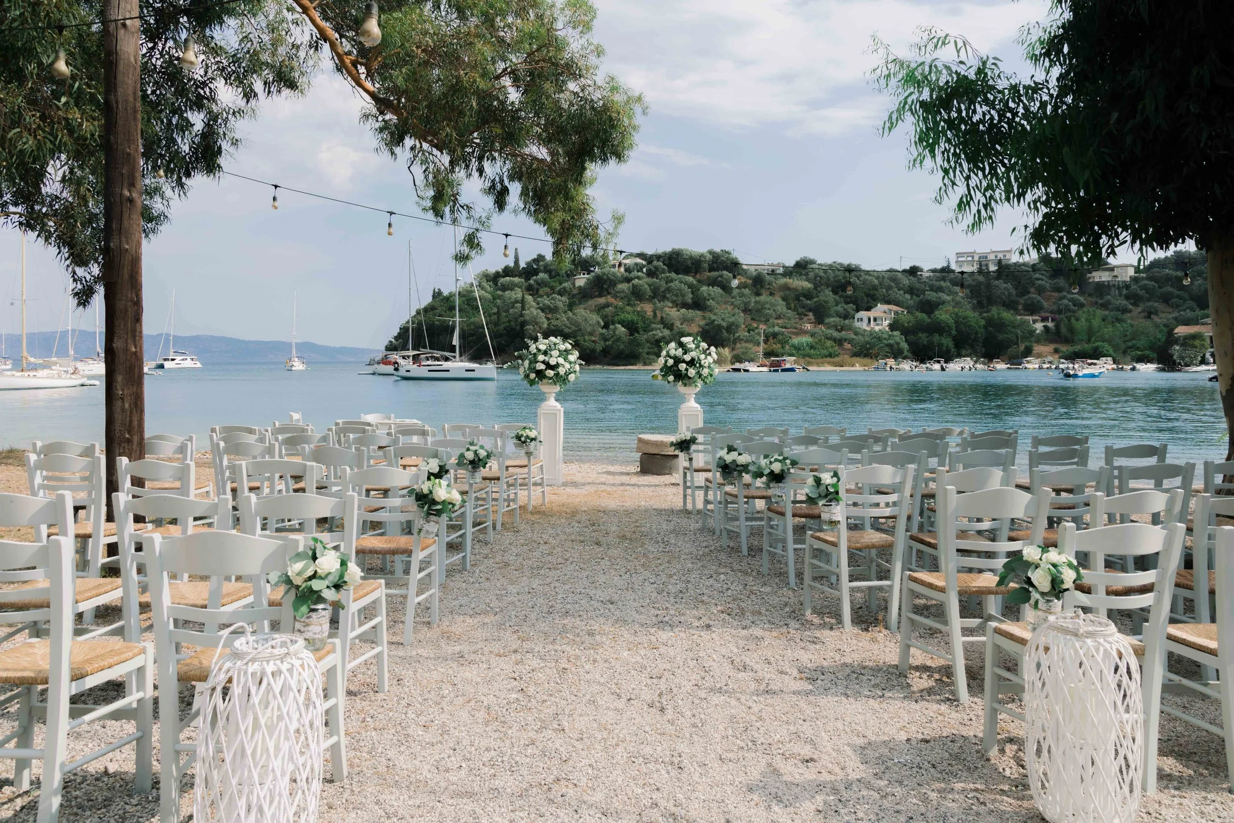 Beachside wedding setup with white chairs decorated with flowers, facing the water with sailboats docked on a calm bay, mountains in the background, and trees surrounding the area.